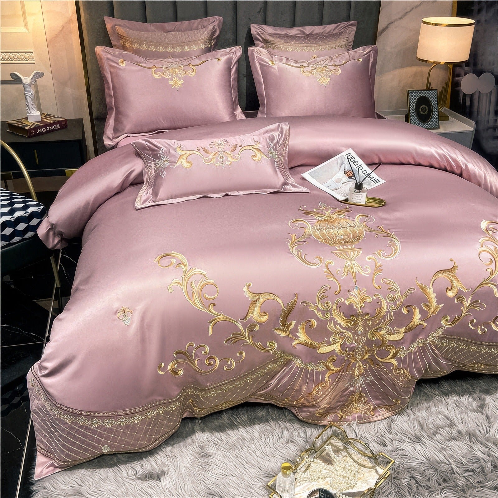 Elegant pink satin bedding set with intricate gold embroidery featuring ornate scroll patterns and a central medallion on the duvet cover. Coordinating pillowcases and decorative cushions are arranged on a plush gray headboard. A gold tray with a magazine and diffuser is placed on the bed, with a cozy white rug beneath.

