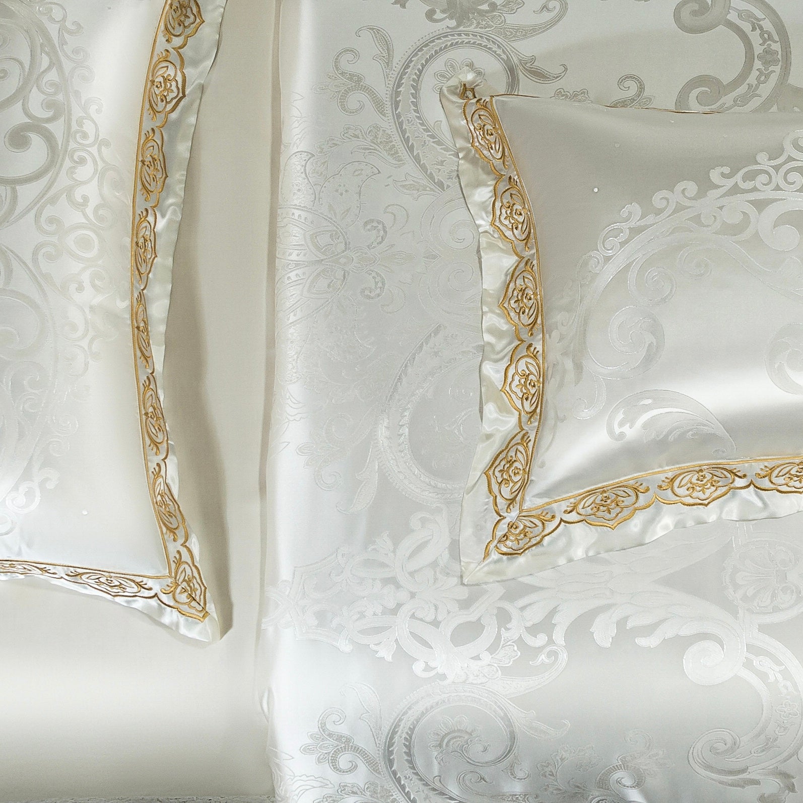 A detailed view of a white jacquard bedding set with intricate swirling patterns and floral motifs woven into the fabric. The edges of the pillowcase and duvet cover are adorned with golden embroidered borders, adding an elegant and regal touch to the design. The smooth and lustrous texture of the fabric enhances its luxurious appeal.

