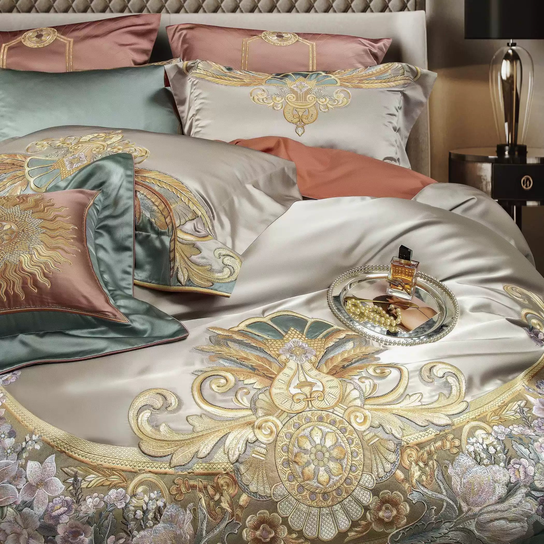 A luxurious bedding set in soft beige satin adorned with intricate golden embroidery featuring floral and geometric motifs. The elegant design is complemented by accent pillows in mint green and blush pink, each with its own ornate detailing, including a radiant sun and intricate scrollwork. A decorative tray with a perfume bottle and pearls adds a touch of sophistication and charm to the opulent display. The quilted headboard and ambient lighting enhance the regal and refined atmosphere of the bedroom sett