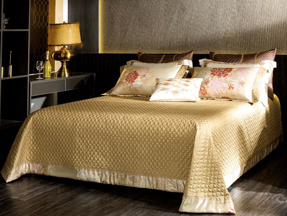 A luxurious bedroom setup featuring a golden quilted bedspread paired with embroidered ivory pillowcases and a matching decorative cushion. The pillowcases are adorned with elegant floral patterns in shades of red, pink, and pastel blue, complemented by a rich golden border with intricate detailing. The warm lighting from a golden lamp enhances the opulent ambiance of the room, which includes a dark wood nightstand and subtle metallic accents for a sophisticated and regal aesthetic.