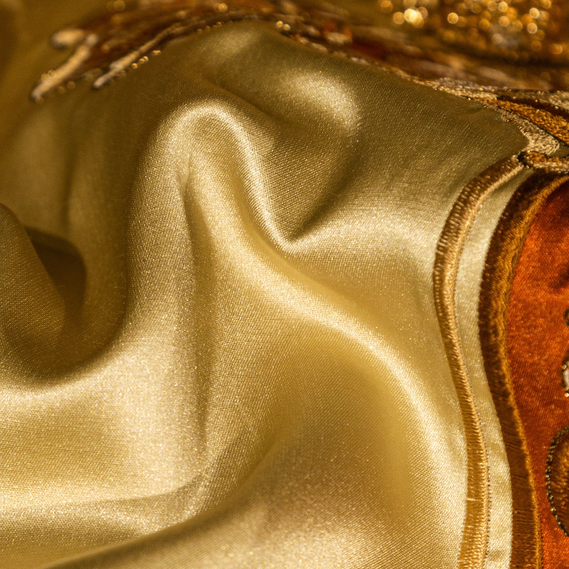 A close-up of shimmering golden satin fabric, displaying its smooth, flowing texture and subtle sheen. The edge is adorned with intricate embroidery in rich orange and gold tones, adding depth and an opulent touch to the luxurious design. The interplay of light highlights the rich quality and elegance of the material.