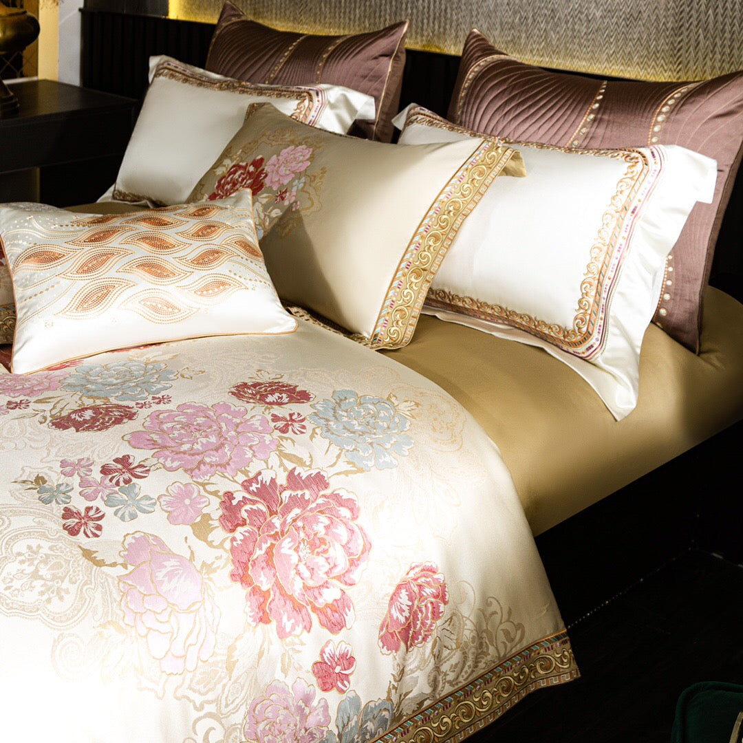 A detailed view of a premium bedding set featuring an ivory duvet cover adorned with intricate floral embroidery in shades of pink, red, and light blue. The borders showcase gold accents and elegant patterns that enhance the luxurious feel. The set is complemented by matching pillowcases with similar designs and golden embroidery, along with quilted mauve decorative pillows in the background. A standout decorative pillow with golden wave-like embellishments adds a touch of sophistication to the ensemble. Th