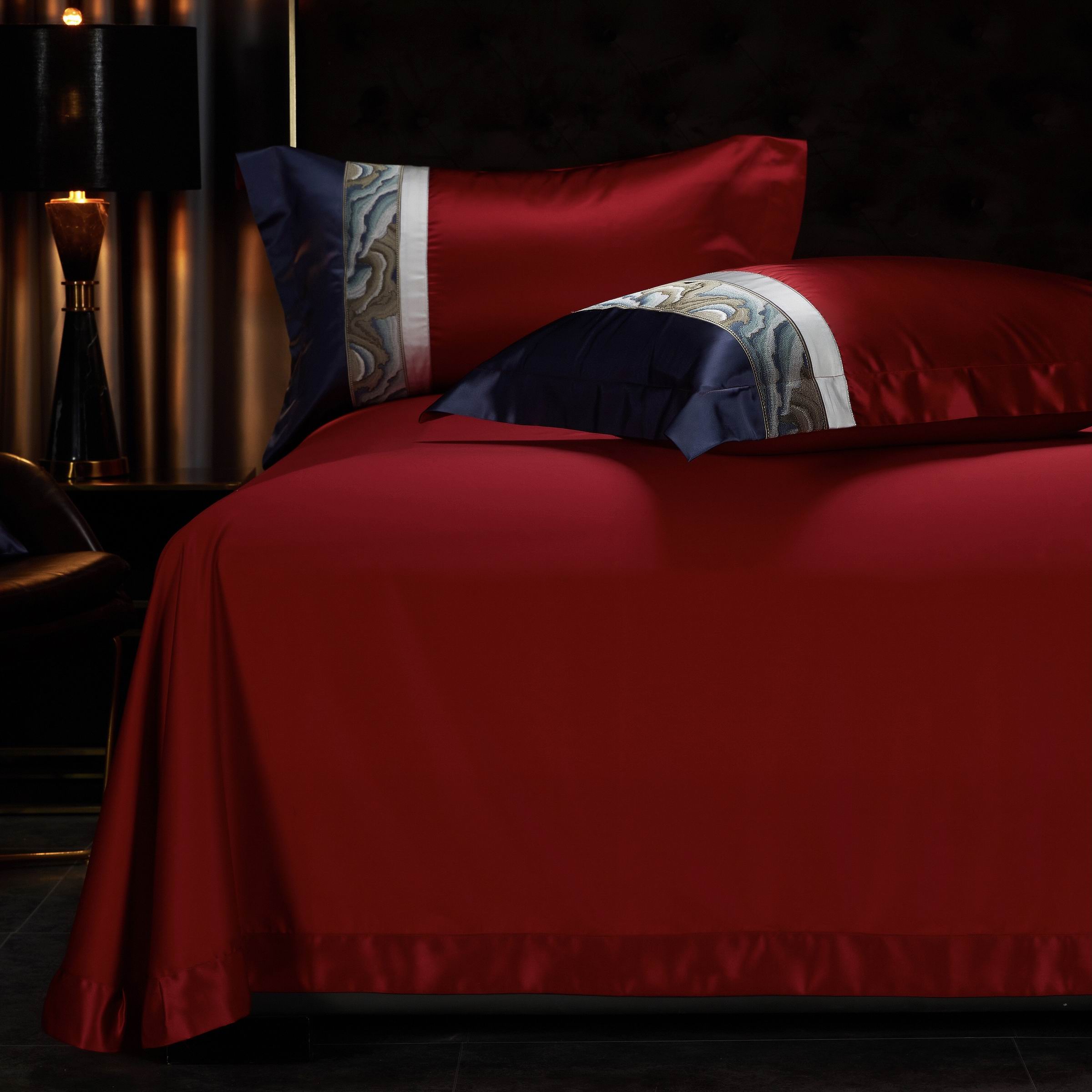 Red bedding with a smooth, satin-like finish, complemented by two pillows featuring navy and white embroidered trims with intricate wave patterns.