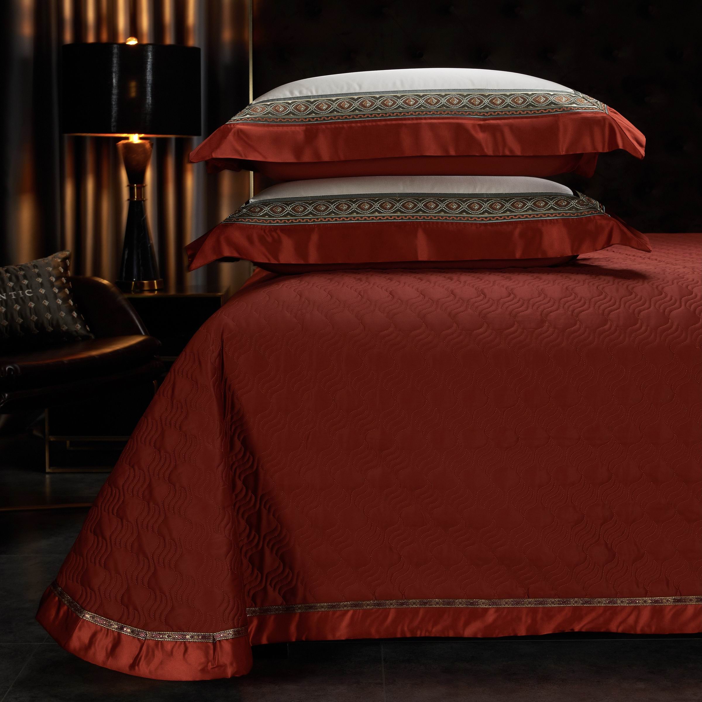 The rich burgundy bedding set features a textured quilt with an elegant wave pattern, exuding a sense of luxury and warmth. The pillowcases showcase intricate geometric embroidery in gold and green hues, bordered with satin trims that highlight their refined craftsmanship. The deep color palette is complemented by soft ambient lighting and a black table lamp with a gold base, creating a cozy yet sophisticated atmosphere. The overall arrangement adds a regal touch to the bedroom, perfectly blending modern de