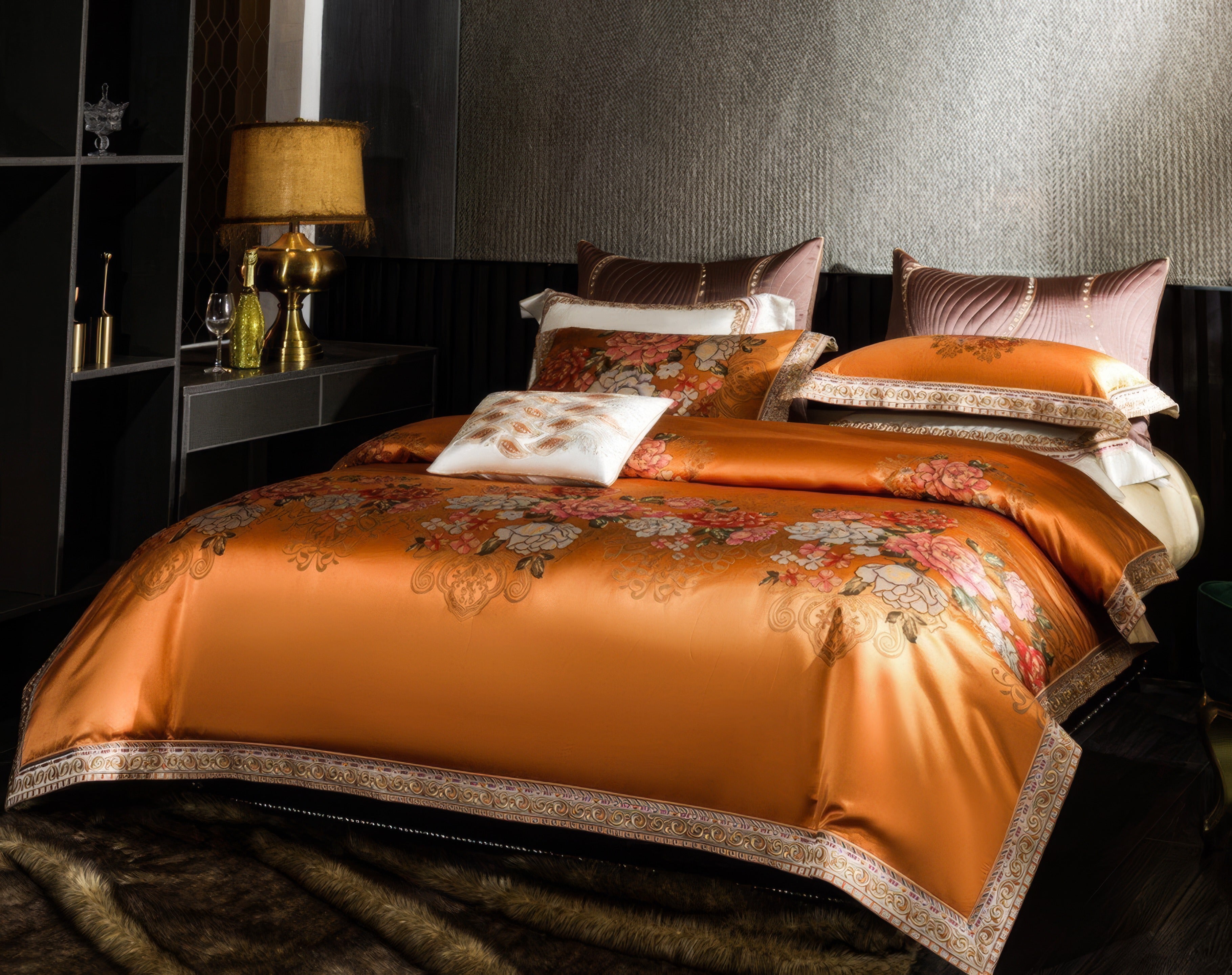 A refined bedroom ensemble showcasing a rich orange bedding set with elaborate floral patterns, highlighted by delicate golden accents and bordered detailing. The duvet cover, along with its matching pillowcases, creates an atmosphere of opulence and warmth. The setting includes a stylish side table adorned with a gold-toned lamp, decorative bottles, and glassware, adding a touch of sophistication. The backdrop features a textured wall complemented by soft lighting, while a fur-style rug enhances the luxuri