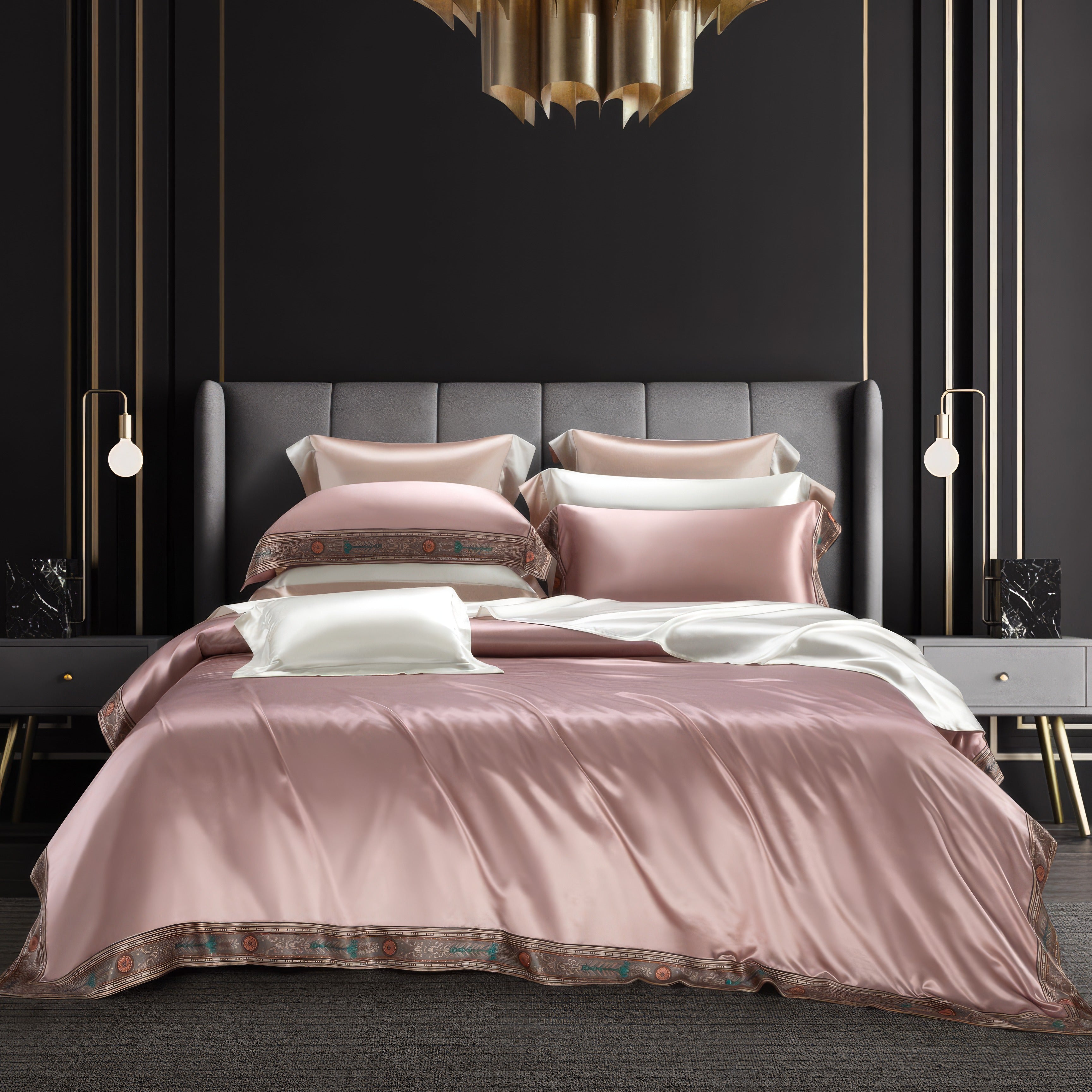 A luxurious bedding set in a soft blush pink hue adorned with intricate patterned borders. The ensemble includes coordinated pillowcases and a neatly draped duvet cover, paired with a pristine white sheet. The sophisticated backdrop features a dark wall with gold accents, complemented by modern pendant lighting and a chic gray headboard, exuding elegance and refinement.