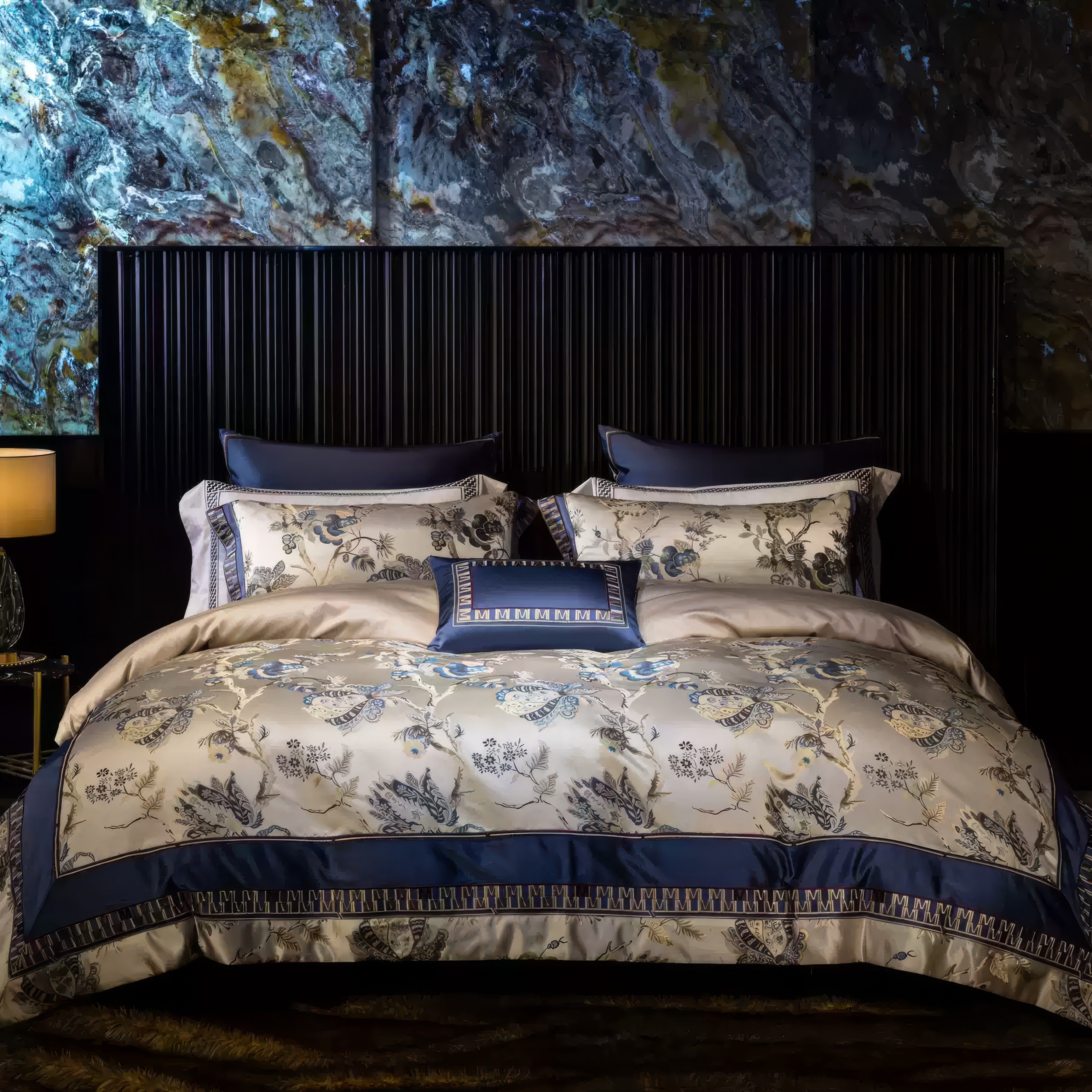 A luxurious bedding set featuring a nature-inspired design with intricate embroidery of blue and ivory botanical motifs on a champagne-colored background. The set includes matching pillowcases with navy blue borders and geometric trim, complemented by a decorative cushion with coordinated embroidery. The bedding is styled against a modern dark wood-paneled headboard with a dramatic marbled accent wall in earthy tones. A warm golden table lamp and a dark side table complete the elegant and opulent bedroom am