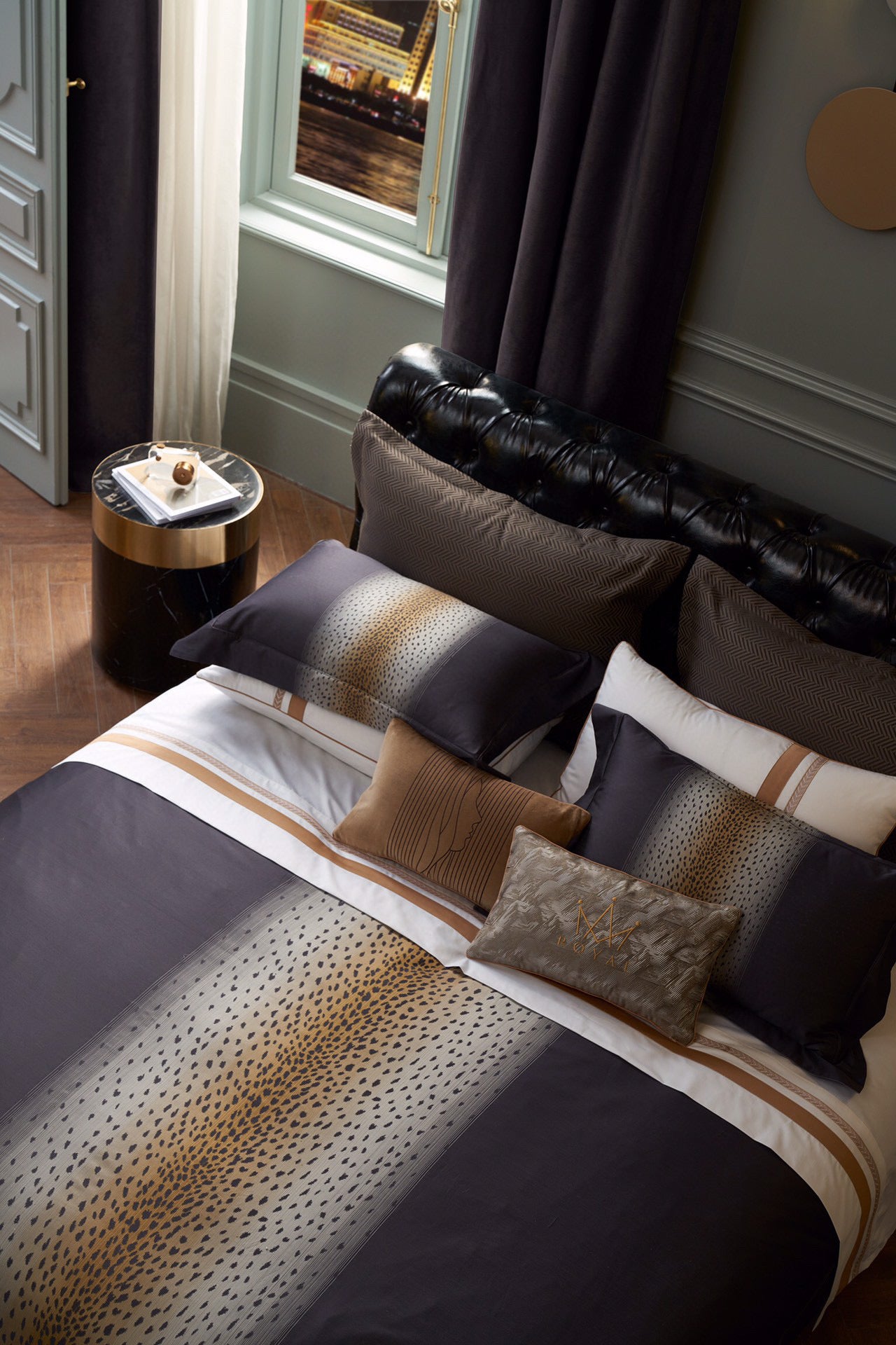 Bedding set in Egyptian cotton featuring a central leopard print gradient design in warm gold and black tones. Paired with coordinating decorative pillows, including embroidered and velvet accents, arranged on a tufted leather headboard. Styled in a sophisticated bedroom with elegant furnishings and a nighttime cityscape visible through large windows.

