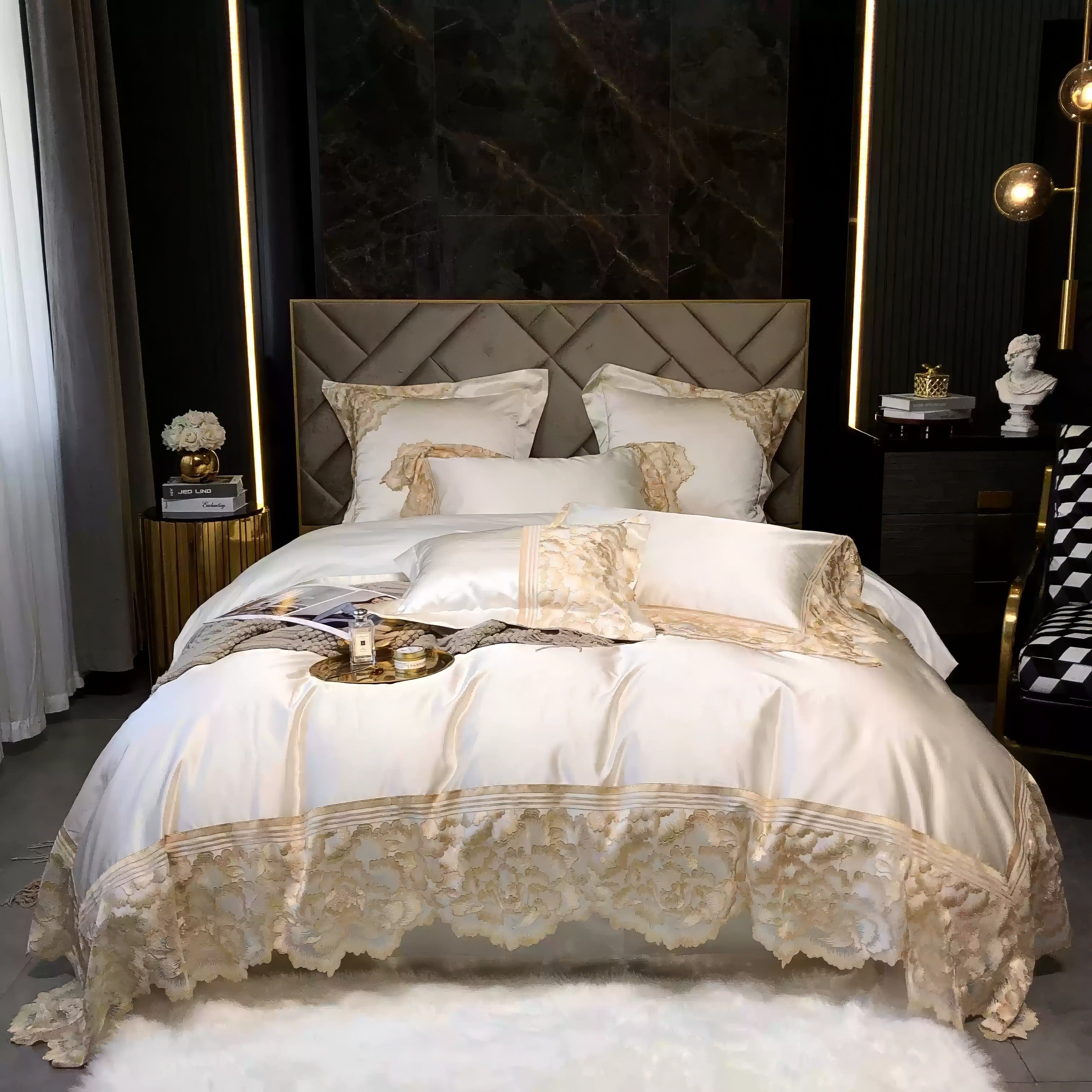 White satin bedding set with gold lace floral trim, neatly arranged pillows, and a matching duvet, styled against a tufted headboard and dark marble wall, with a soft white rug below.

