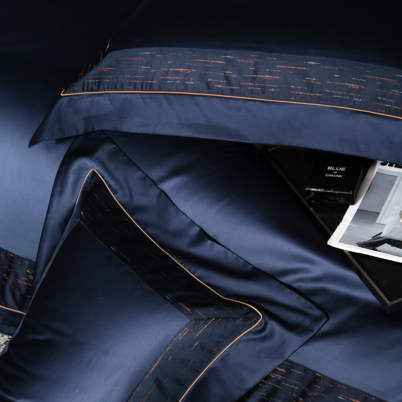 Elegant navy blue bedding with copper and silver accents, showcasing a close-up of a decorative pillow and duvet. The luxurious fabric features refined stitching and a subtle sheen, enhanced by a modern streak pattern. Stylish details, including orange piping, add a contemporary and sophisticated touch.

