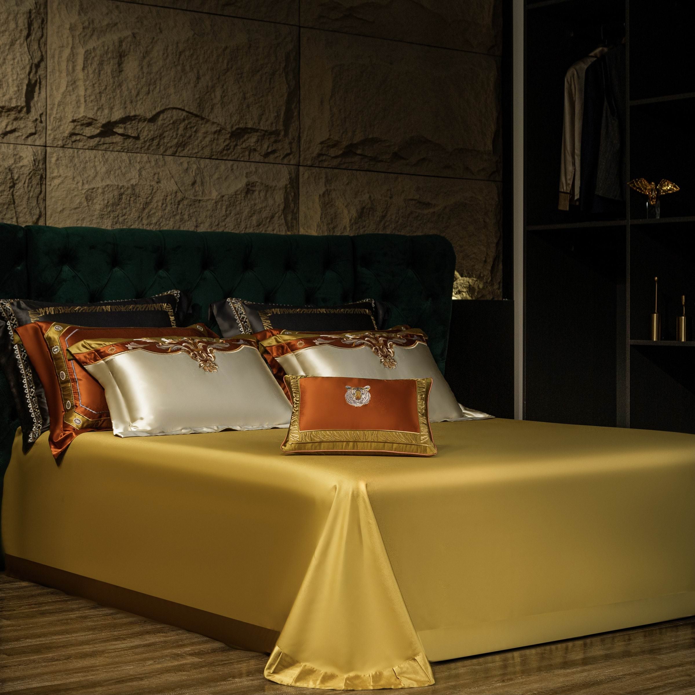 A luxurious bedroom setting featuring a gold satin bedspread that elegantly drapes to the floor, complementing the intricate design of the pillows. The decorative pillows display rich gold, ivory, and bronze tones with ornate embroidery, highlighted by an emerald green tufted headboard that adds depth and sophistication. The textured stone wall and ambient lighting enhance the opulent atmosphere, creating a regal and refined aesthetic.