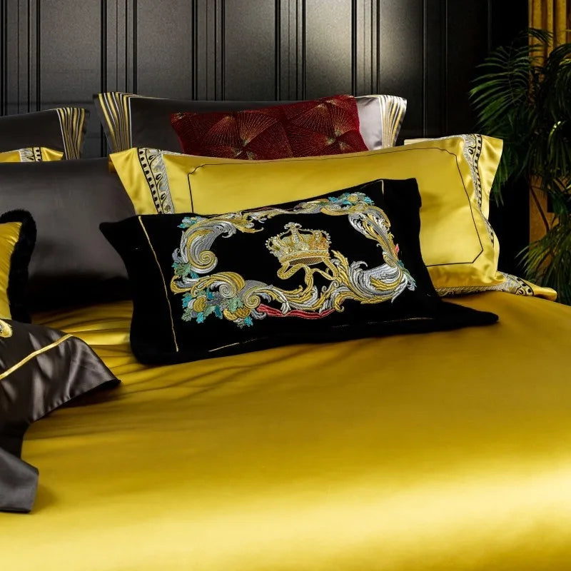 A detailed shot of a luxurious bedding set highlighting a black velvet throw pillow adorned with intricate multicolored embroidery, featuring a golden crown motif surrounded by ornate patterns. Behind it, a bold yellow pillow with black piping complements the design, accompanied by a textured red pillow in the background. The rich yellow satin fabric of the duvet creates a vibrant and regal aesthetic, enhanced by the elegant interplay of colors and textures.

