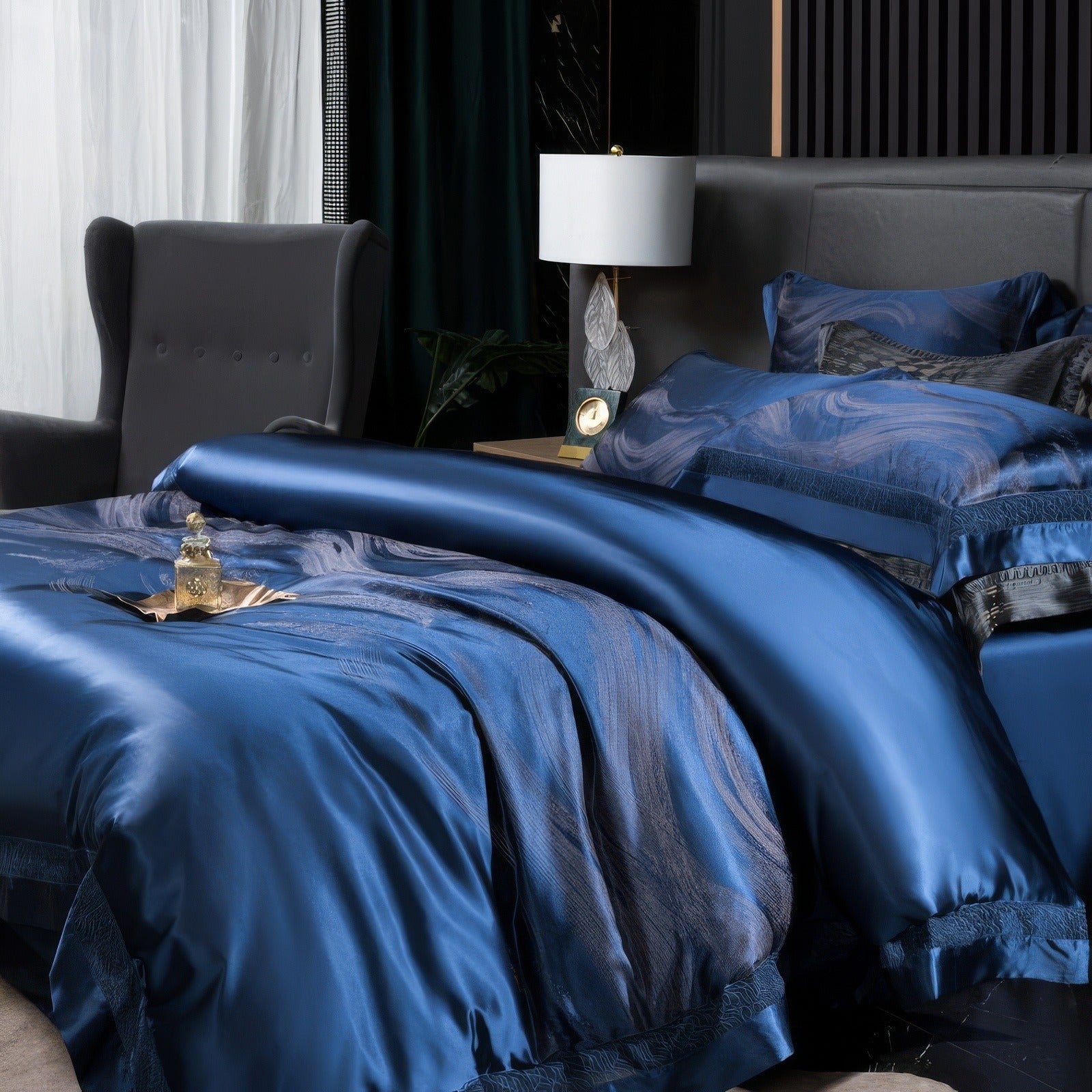 A luxurious navy blue bedding set with a silky finish and intricate subtle patterns lies elegantly on a bed. The bedding reflects a sheen that highlights its premium quality. Beside the bed, a modern bedside table with a decorative lamp enhances the sophisticated ambiance. A small ornate golden tray with a perfume bottle rests atop the comforter, adding a touch of elegance to the rich setting. The wingback chair in the background completes the cozy, upscale look.

