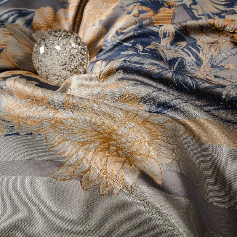 Close-up of a dark gray jacquard fabric adorned with intricate golden floral designs and subtle blue accents. A shimmering crystal orb is nestled within the tufted center, enhancing the luxurious texture and detailed craftsmanship of the bedding material.