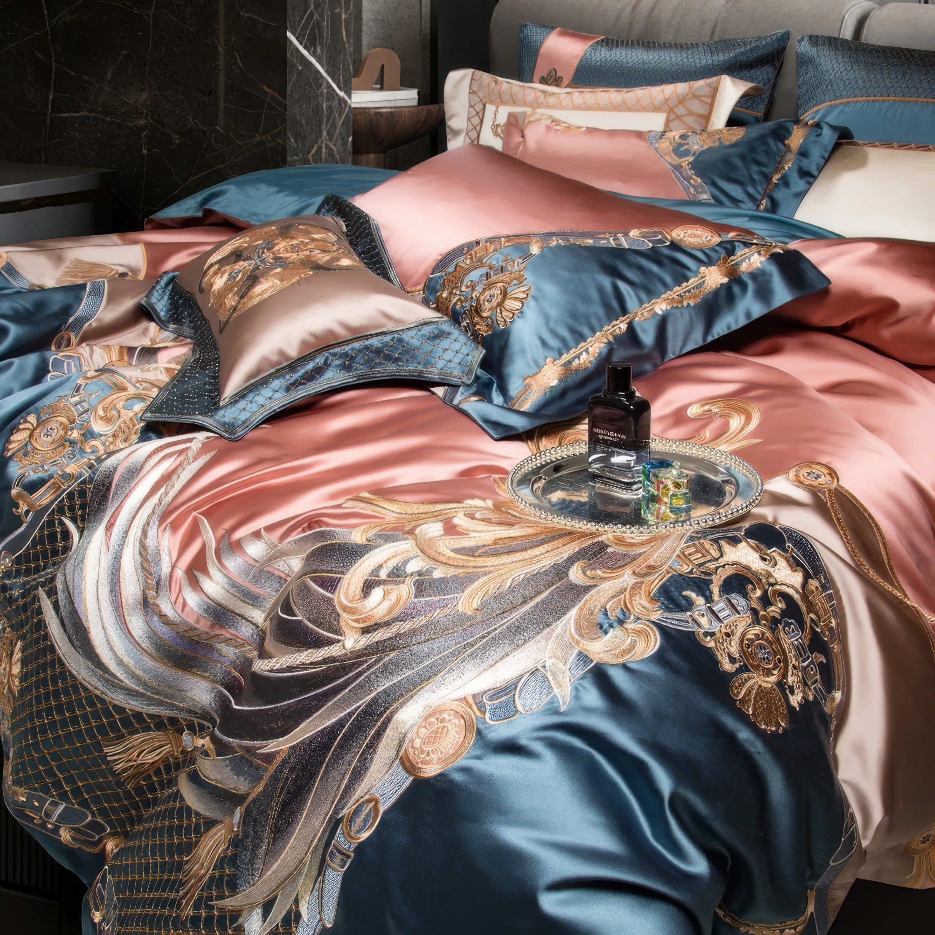 Luxurious bedding set featuring intricate golden baroque patterns on satin fabric in shades of rose gold and teal. The close-up highlights the artistic details of the duvet cover, with swirling metallic designs and ornate accents. Coordinating pillows with embroidered patterns add texture and elegance. A silver tray with a perfume bottle and colorful accents enhances the sophisticated ambiance. The background reveals a marble wall and plush headboard, completing the opulent, modern setting.