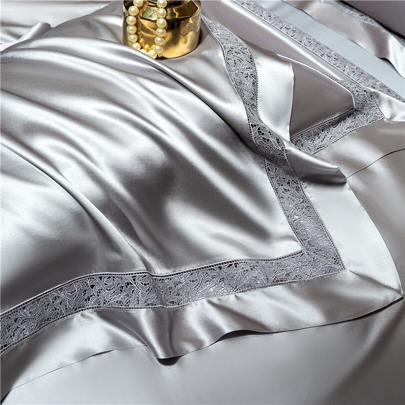 Luxurious silver satin bedding with intricate lace borders draped elegantly. The smooth, lustrous fabric reflects the soft lighting, creating a shimmering effect. A golden decorative vase with pearl beads adds a touch of opulence to the composition.