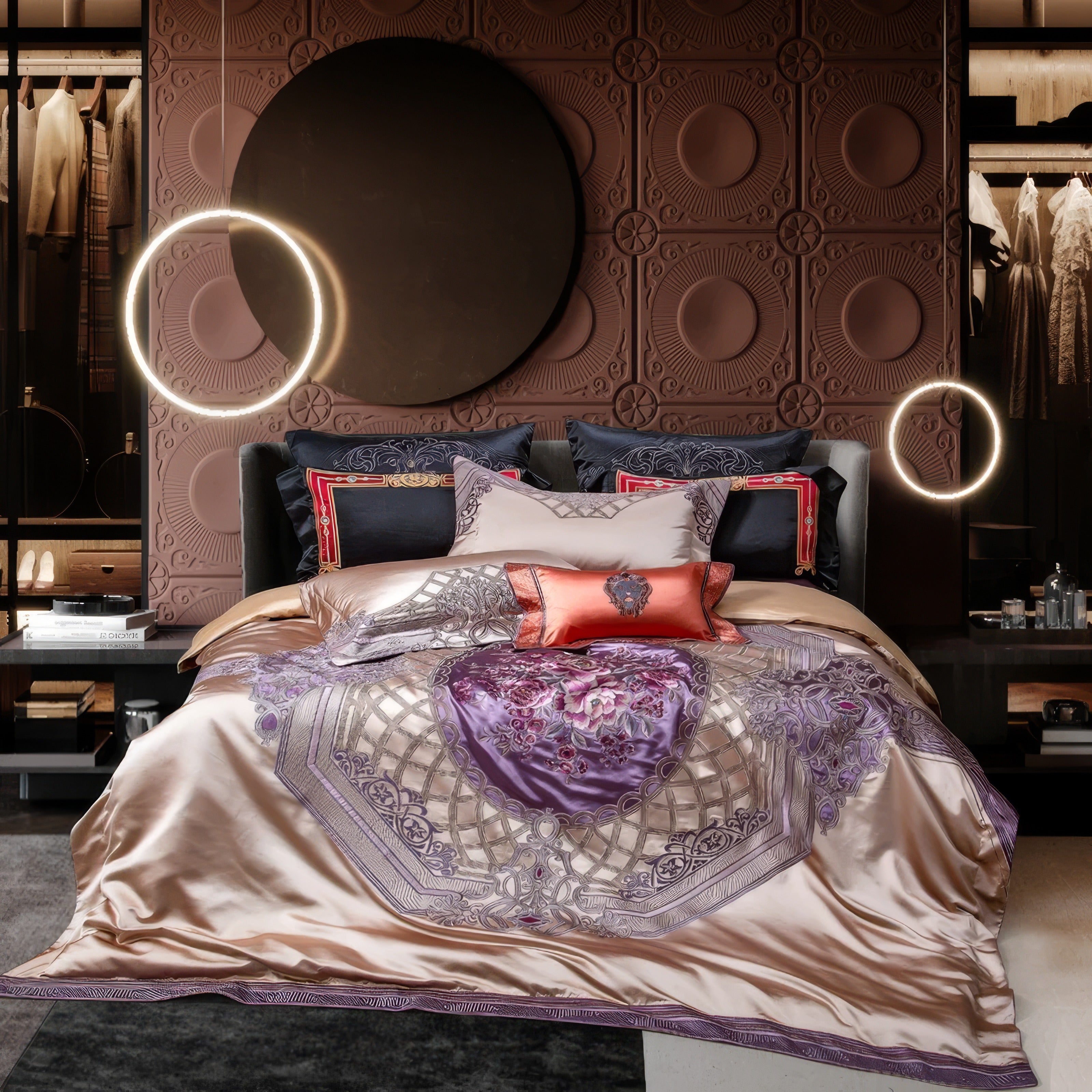 A luxurious bedding arrangement featuring a silky champagne-colored duvet cover with intricate floral embroidery in purple and silver tones. The design is complemented by matching pillowcases in soft pastels and accent pillows in vibrant coral and navy blue with regal patterns. The backdrop includes a textured, decorative wall panel in a warm brown hue, adorned with a large black circular mirror. Two modern ring lights add a contemporary touch, illuminating the sophisticated setting. A walk-in closet with n