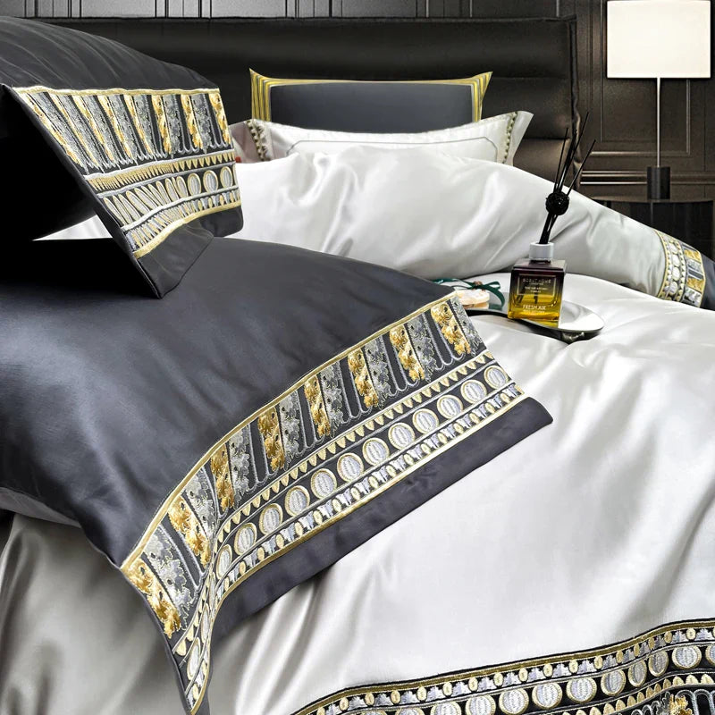 A detailed close-up of a luxury bedding set featuring black satin pillowcases with intricate golden and silver embroidered patterns along the borders. The embroidery showcases ornate designs with metallic accents, adding an opulent touch. The pillowcase rests against a pristine white duvet, accented with matching gold and silver embroidery. A stylish diffuser bottle is placed on the bed, enhancing the luxurious ambiance. 