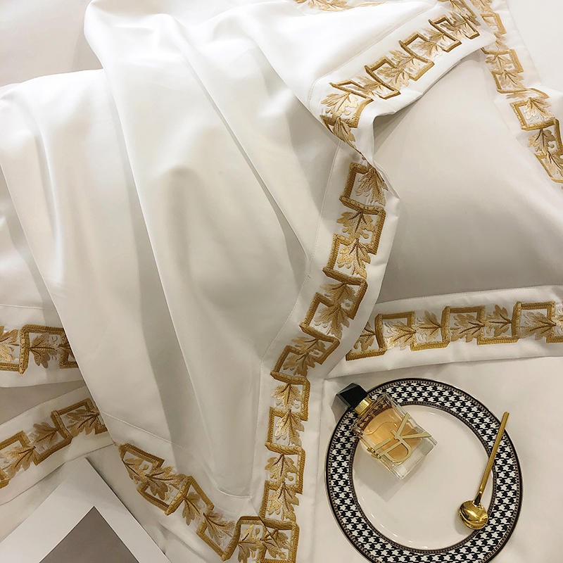 Egyptian cotton bedding in white with intricate golden embroidery, styled alongside a patterned plate, gold spoon, and luxury perfume for an opulent presentation.

