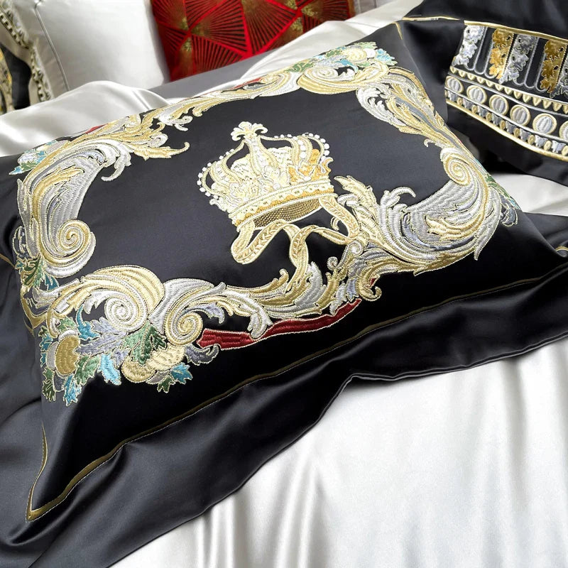 A close-up view of an ornate decorative pillow featuring intricate embroidery on a black satin base. The design highlights a golden crown surrounded by elegant swirls, floral accents in gold, silver, red, and green, and intricate detailing, exuding a regal and luxurious charm. The soft, glossy texture of the fabric complements the embroidery, while the edges are finely stitched for a polished finish.

