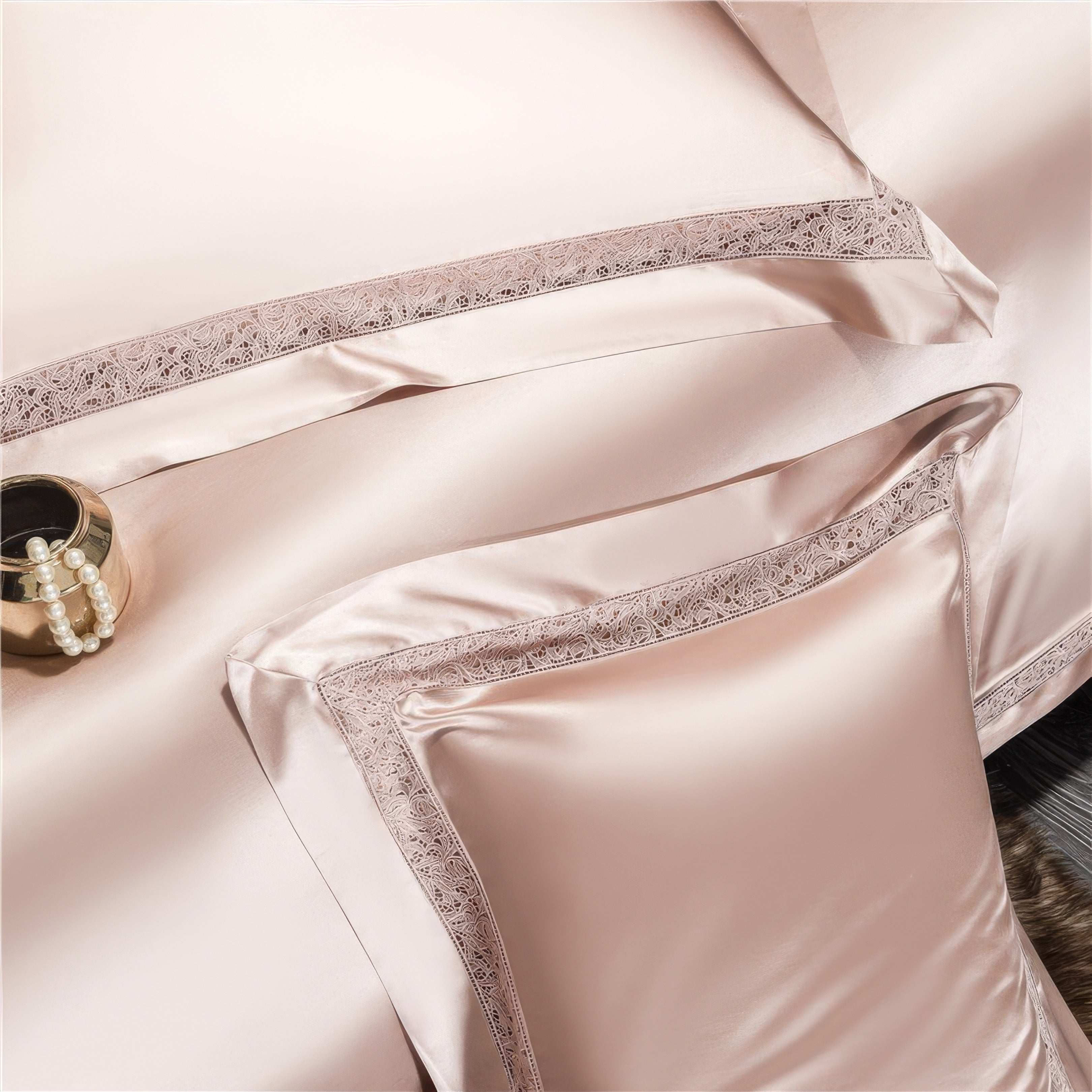 Close-up view of a luxurious champagne-colored satin bedding set featuring pillowcases and a duvet with intricate lace trim detailing. A gold decorative item with pearl accents adds a sophisticated touch, complementing the soft sheen of the fabric and creating an elegant, refined look.
