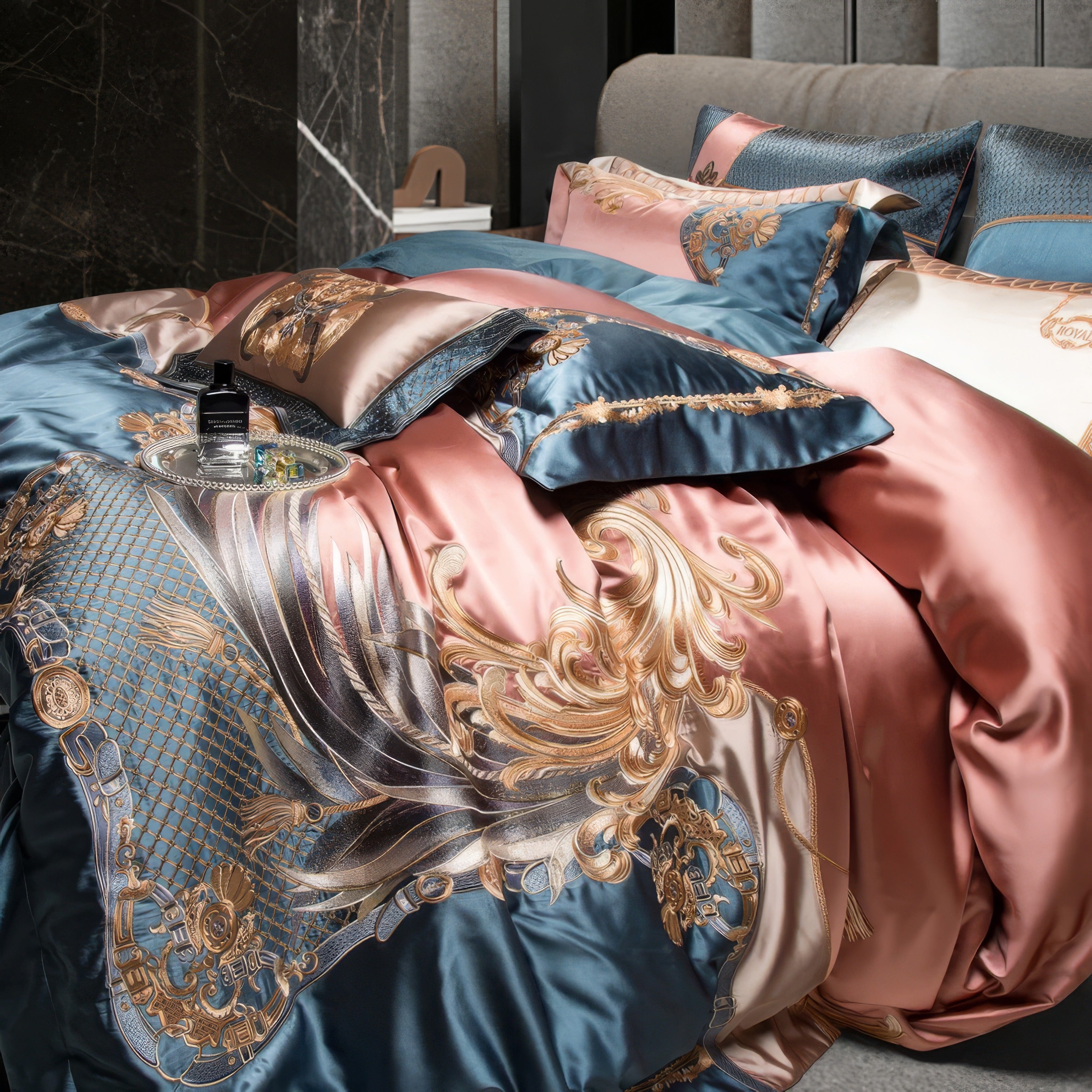 Close-up of a luxurious bedding set adorned with elaborate golden baroque designs on a blend of rose gold and teal satin fabric. The duvet cover showcases intricate swirling patterns with metallic accents, enhancing the opulent feel. Matching pillowcases feature complementary embroidery, adding texture and depth. A glass tray with elegant perfume bottles and accessories sits on the bed, emphasizing a sophisticated lifestyle. The background includes a marble wall and upholstered headboard for a modern, elega