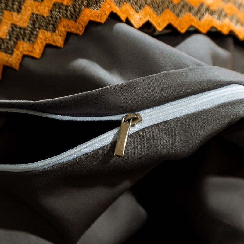 Close-up of a dark gray fabric with a white zipper partially opened, revealing its gold metal zipper pull. Above, an orange and black chevron-patterned trim decorates the fabric, adding contrast to the smooth material.

