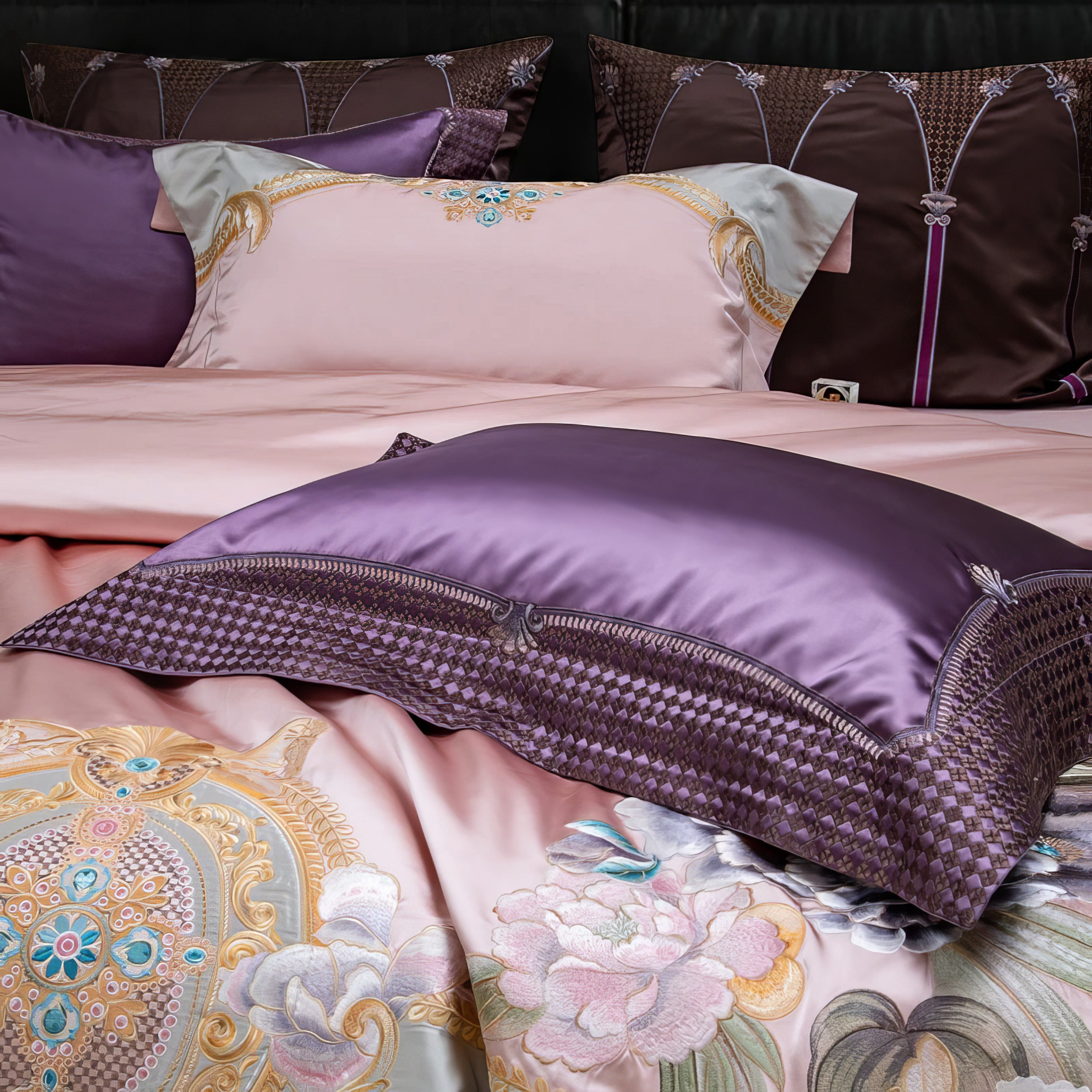 A detailed close-up of a luxury bedding set, featuring a plush purple satin pillow with intricate textured edging, adding depth and elegance. The bedding showcases a soft blush pink base with ornate gold and pastel floral embroidery, complemented by rich plum and brown decorative pillows in the background, creating a sophisticated and opulent bedroom look.