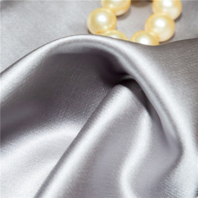 Close-up of smooth silver satin fabric, highlighting its soft, glossy texture and elegant drape. A string of luminous pearls rests gently on the fabric, adding a touch of luxury and sophistication.