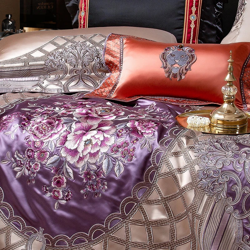 A luxurious close-up of a bedding set showcasing a stunning lavender satin centerpiece adorned with intricate floral embroidery in shades of pink and purple. The detailed embroidery is surrounded by a geometric champagne satin pattern. An orange satin decorative pillow with a regal lion motif enhances the elegance of the arrangement. On the side, a gold tray with a pearl accessory and an ornate golden ornament completes the opulent presentation. The ensemble exudes a sense of royal sophistication and artist