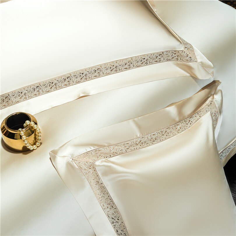 Close-up of luxurious cream-colored pillowcases and a duvet cover, showcasing intricate lace embroidery along the edges. The silky fabric gleams softly under the light, emphasizing its high-quality texture. A gold-accented decorative bowl with pearl accessories adds a touch of sophistication and elegance to the composition.