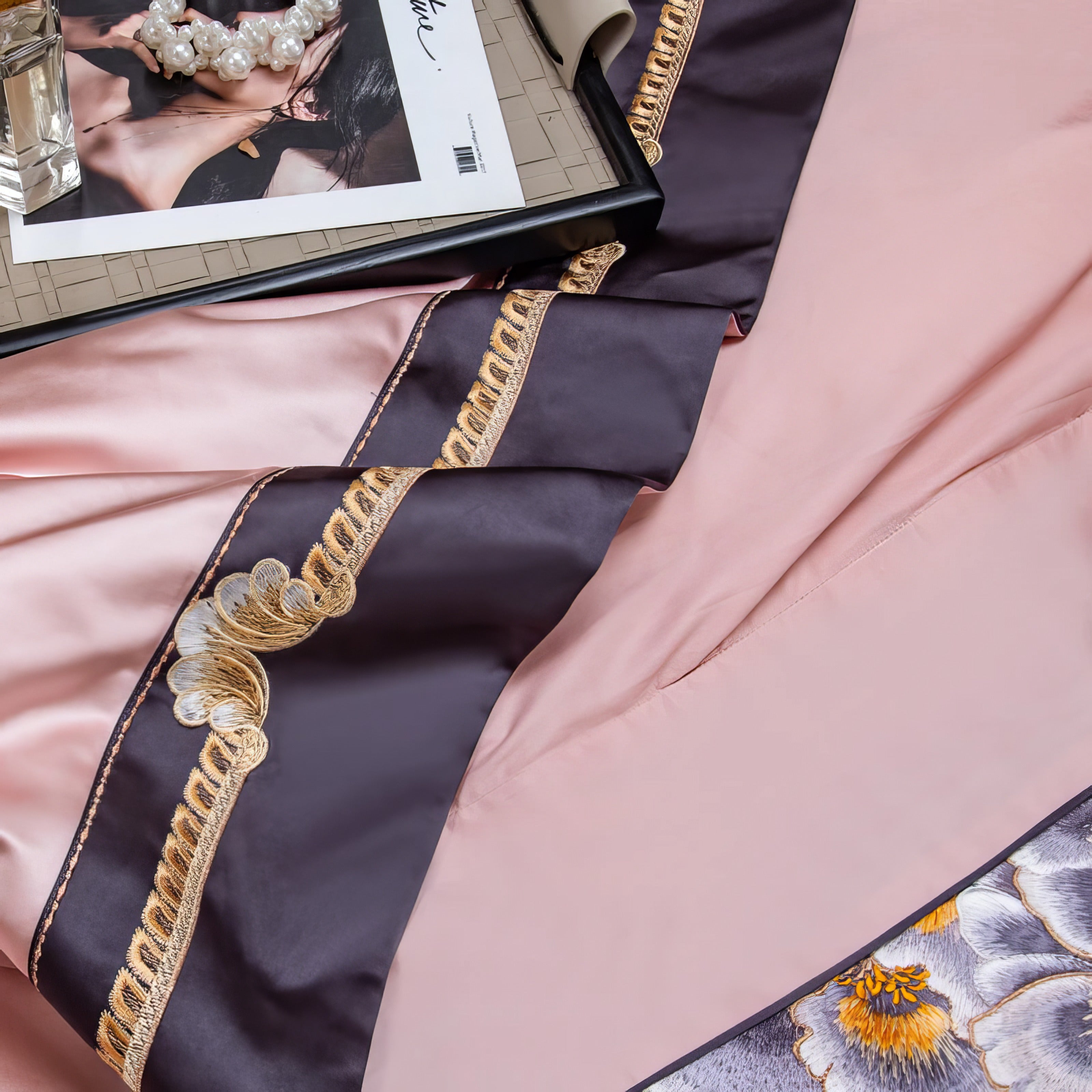 A detailed close-up of a luxurious bedding set showcasing a soft pink duvet with dark brown accents, highlighted by intricate gold embroidery. The delicate floral patterns add an elegant touch, while the silky fabric exudes sophistication and premium quality. A tray with decorative pearls and a magazine enhances the setting's opulent feel.