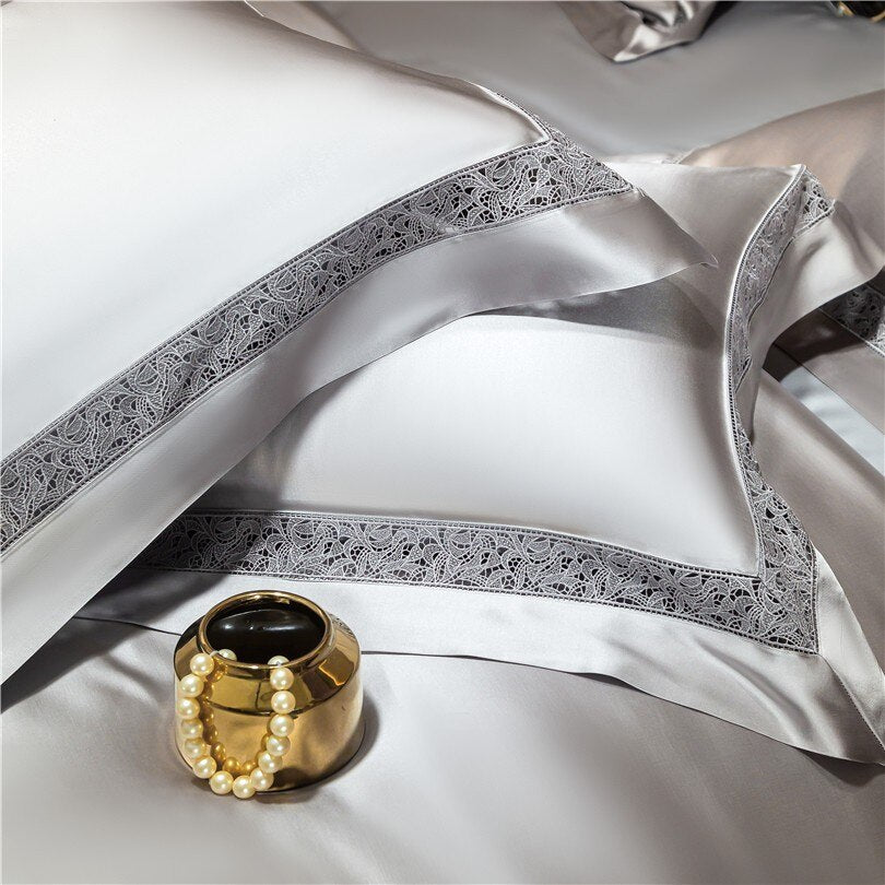 Close-up of silver satin bedding showcasing pillowcases and duvet covers adorned with intricate lace borders. The sleek, shiny fabric emphasizes luxury, complemented by a golden decorative bowl holding a pearl bracelet, adding a touch of sophistication.