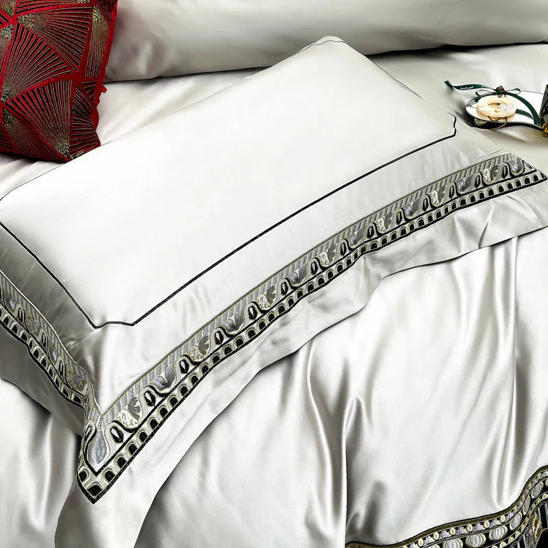 A close-up of a luxurious white pillowcase with a delicate black trim and an ornate embroidered border featuring intricate silver and black patterns. The embroidery adds a regal touch to the sleek satin fabric, showcasing a blend of elegance and sophistication. A vibrant red accent pillow with geometric designs is visible in the background, contrasting beautifully with the white and metallic tones. The bedding exudes a sense of opulence, ideal for a refined bedroom setting.

