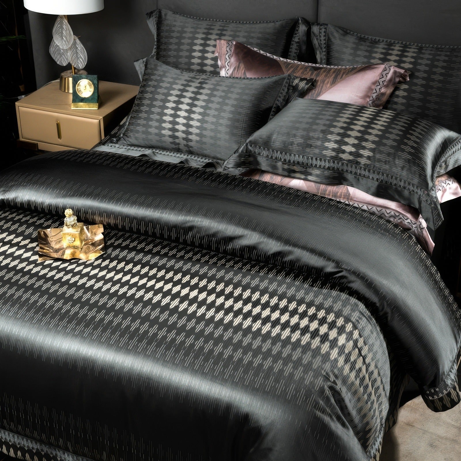 Close-up of a luxurious black bedding set with intricate geometric patterns, accented by soft pink pillows, draped on a contemporary bed with a gold nightstand and decorative perfume tray.

