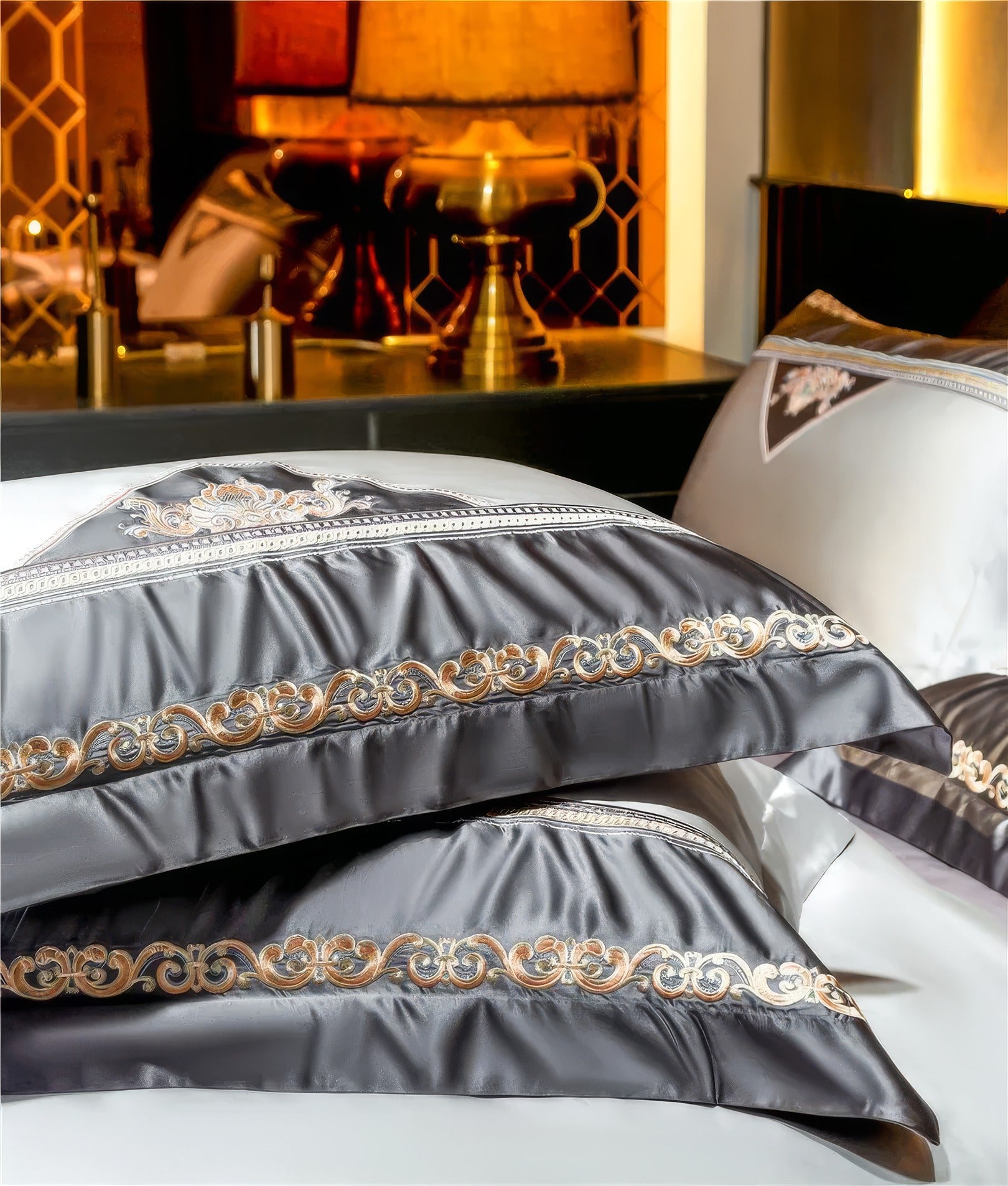 A close-up view of two elegant silver satin pillows adorned with gold and black embroidery in intricate swirling patterns. The luxurious sheen of the fabric highlights the fine craftsmanship. In the background, warm lighting and gold accents on decorative items create a cozy, sophisticated ambiance.

