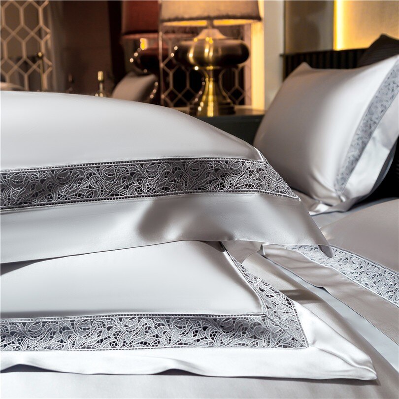 Close-up of silver satin pillowcases stacked neatly, featuring intricate lace borders for a touch of elegance. The smooth, glossy fabric enhances the luxurious feel, with soft lighting and a golden table lamp in the background creating a sophisticated ambiance.