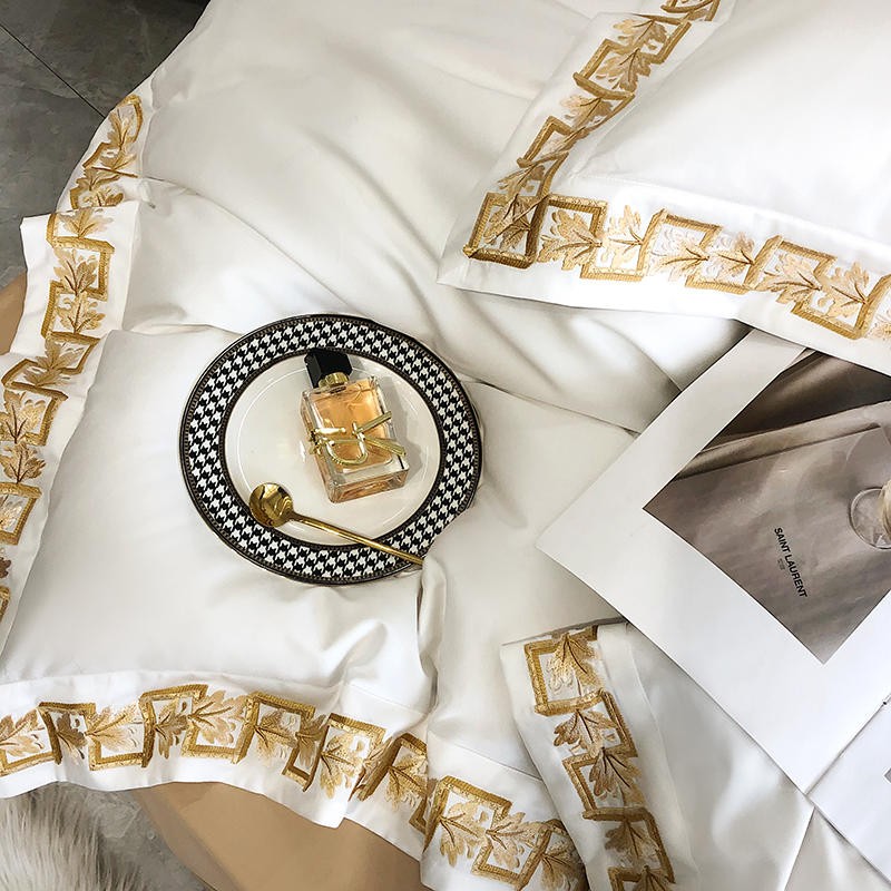 White Egyptian cotton bedding with ornate gold embroidery, styled elegantly with a designer perfume bottle, a houndstooth-patterned plate, a gold spoon, and an open luxury magazine.

