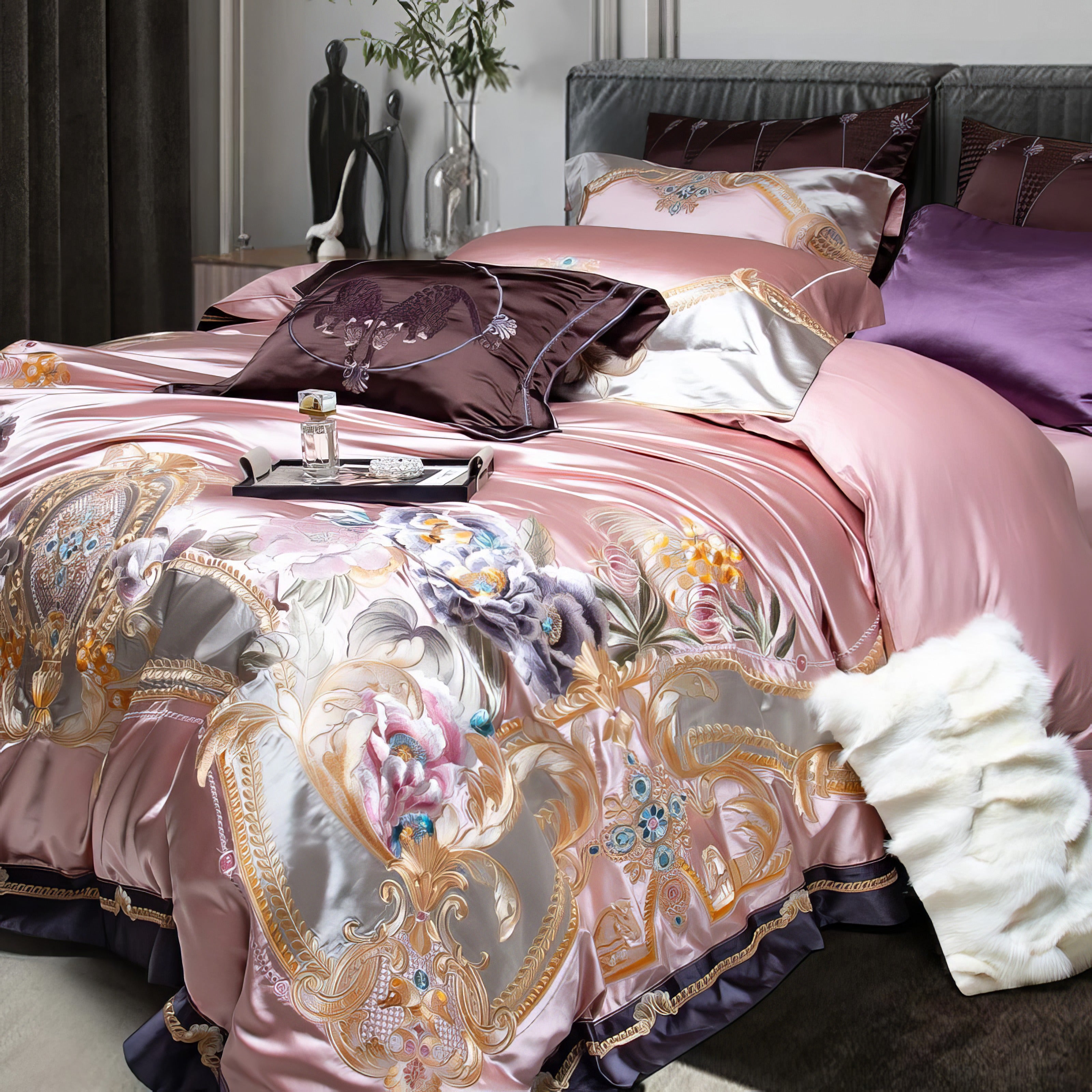 A close-up of an opulent bedding set in blush pink with intricate floral and ornamental embroidery in gold and pastel tones. The silky texture reflects light elegantly, complemented by deep plum and lavender accent pillows. A plush white faux fur throw adds a luxurious touch, enhancing the cozy yet regal ambiance of the setting.