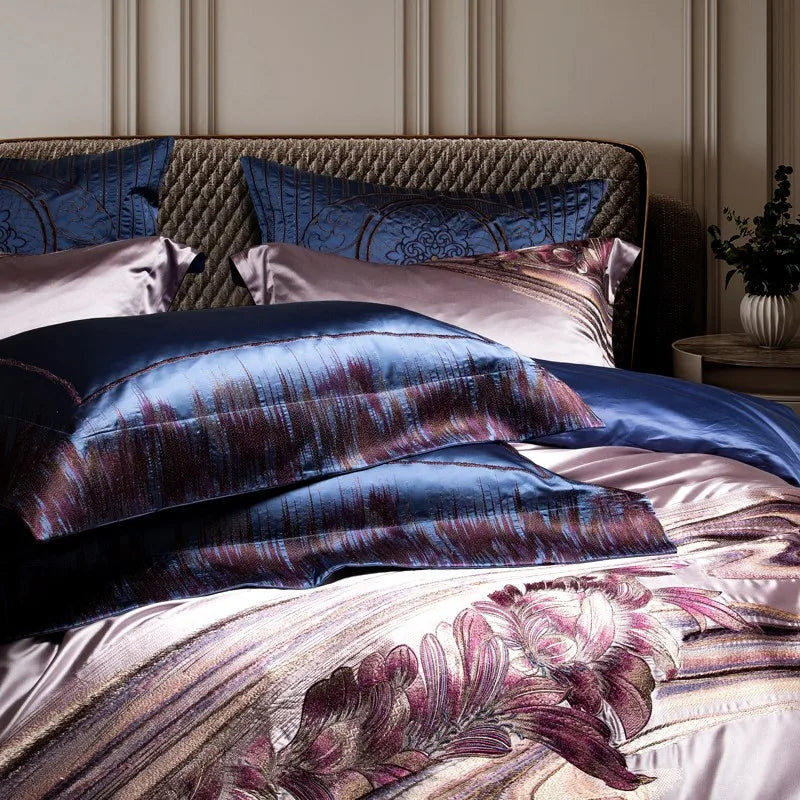 A layered arrangement of bedding featuring royal blue silk pillowcases adorned with shimmering plum and burgundy embroidery, complementing lavender silk fabric with pink floral detailing. The quilted headboard and additional embroidered pillows in the background add depth and sophistication.