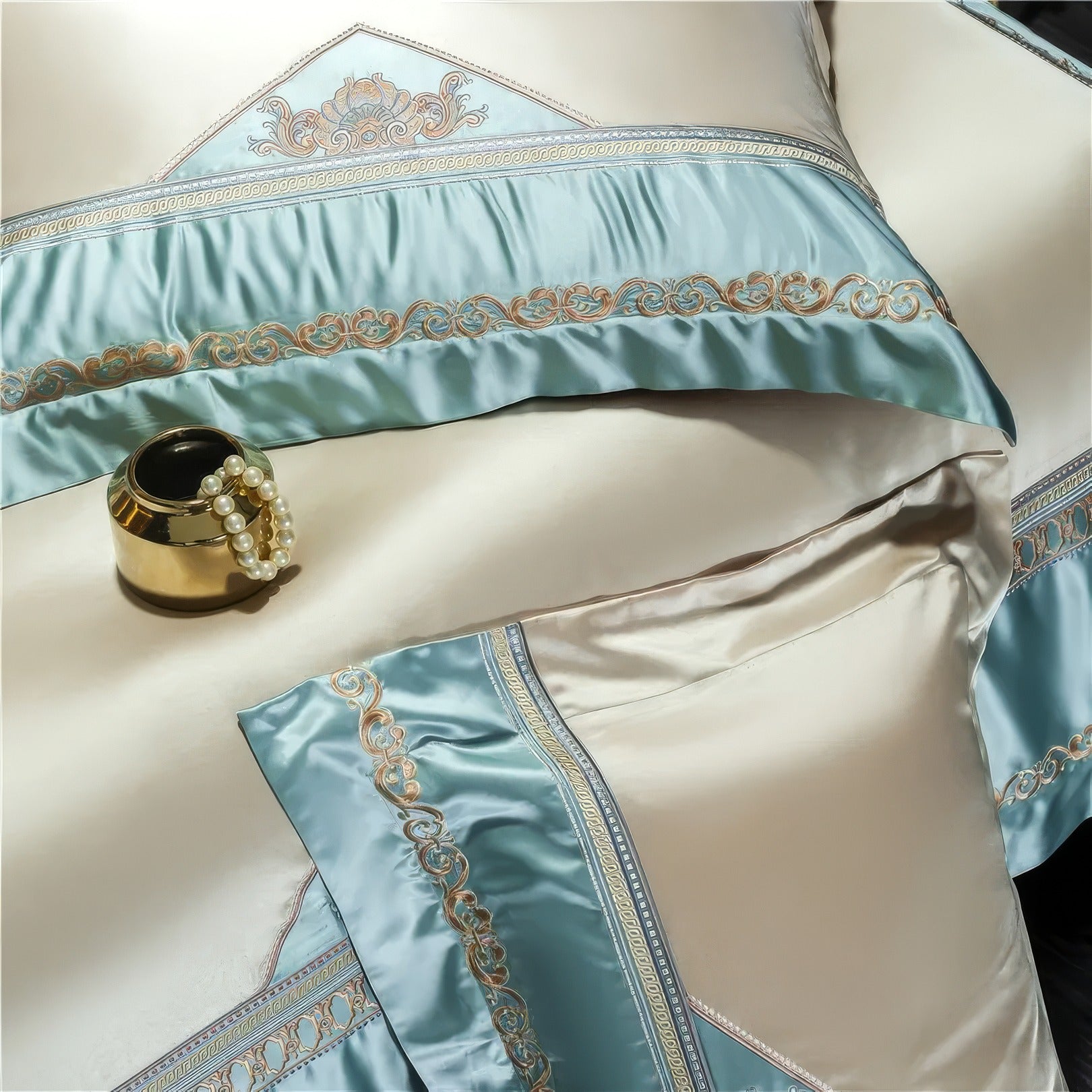 A close-up view of a luxurious beige and teal bedding set, featuring intricate gold embroidery on the teal accents. A gold decorative jar with a string of pearls sits elegantly atop the bedding, highlighting its opulence.

