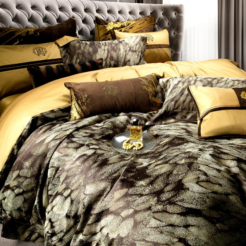 A luxurious bedding set featuring a rich combination of brown and gold tones with a textured feather-inspired pattern. The set includes decorative pillows, embroidered cases, and elegant golden accents. The tufted headboard in a soft grey fabric adds a touch of sophistication, while the silver tray with a golden perfume bottle and pearl necklace on the bed completes the opulent look. This setup embodies elegance and high-end design, ideal for luxury interior styling or bedding collection advertisements.