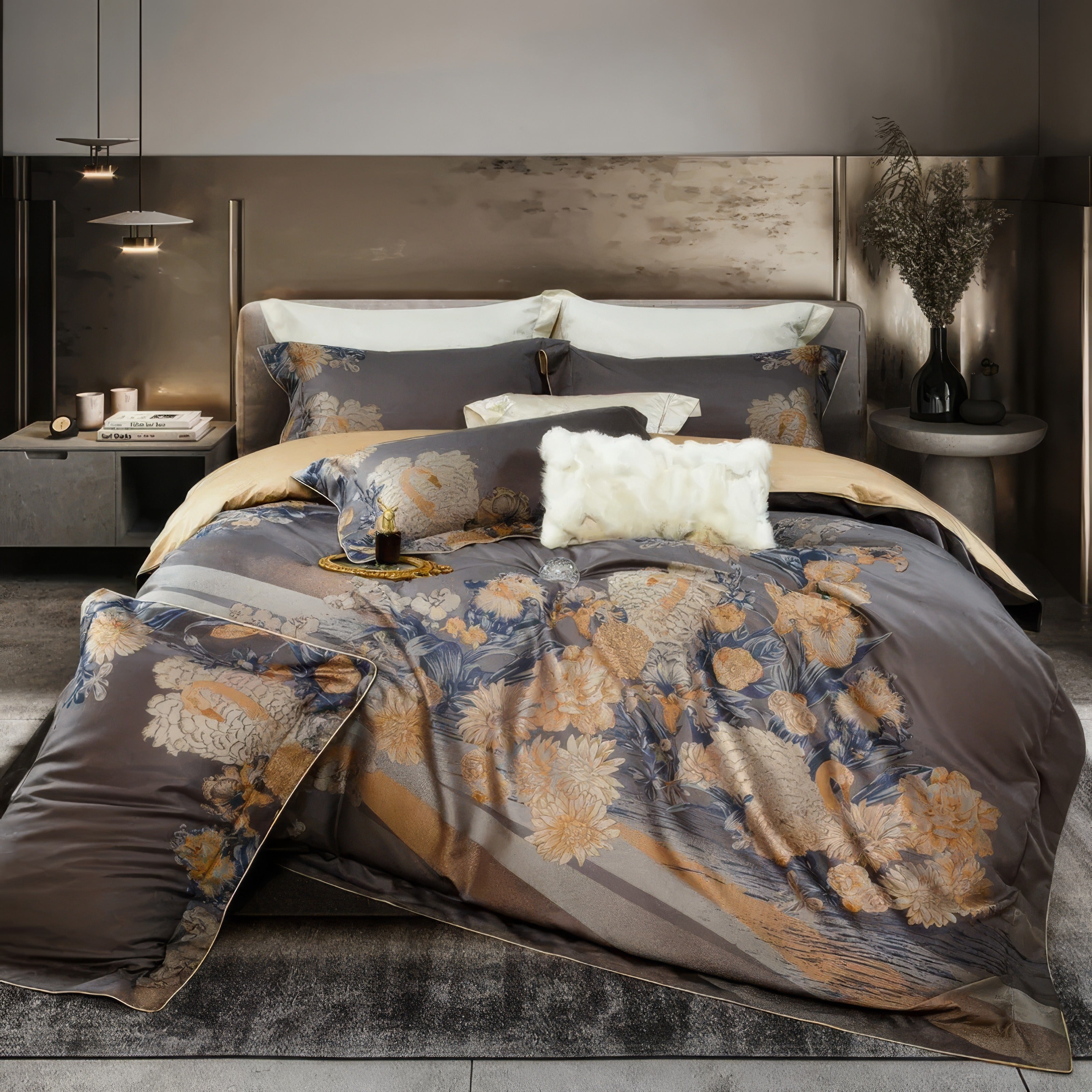 A luxurious bedding set featuring a charcoal-gray duvet adorned with intricate floral patterns in shades of gold and blue. Matching pillowcases complement the design, while cream-colored accent pillows and a fluffy white decorative pillow add layers of texture and contrast. The bed is framed by a sleek metallic headboard, with modern pendant lights and minimalistic side tables enhancing the sophisticated atmosphere of the room.