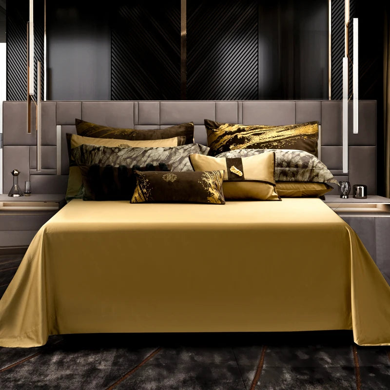 A full view of a luxurious bed setting featuring a sleek gold bedspread that flows elegantly to the floor. The bed is adorned with multiple decorative pillows in varying designs and textures, including velvet pillows with golden splashes, patterned brown and gold pillows, and rich earth-tone accents. The headboard showcases a modern geometric design in soft gray, complemented by a dramatic black textured wall behind it. Hanging lights and bedside tables with minimalist decor add a contemporary touch to the 