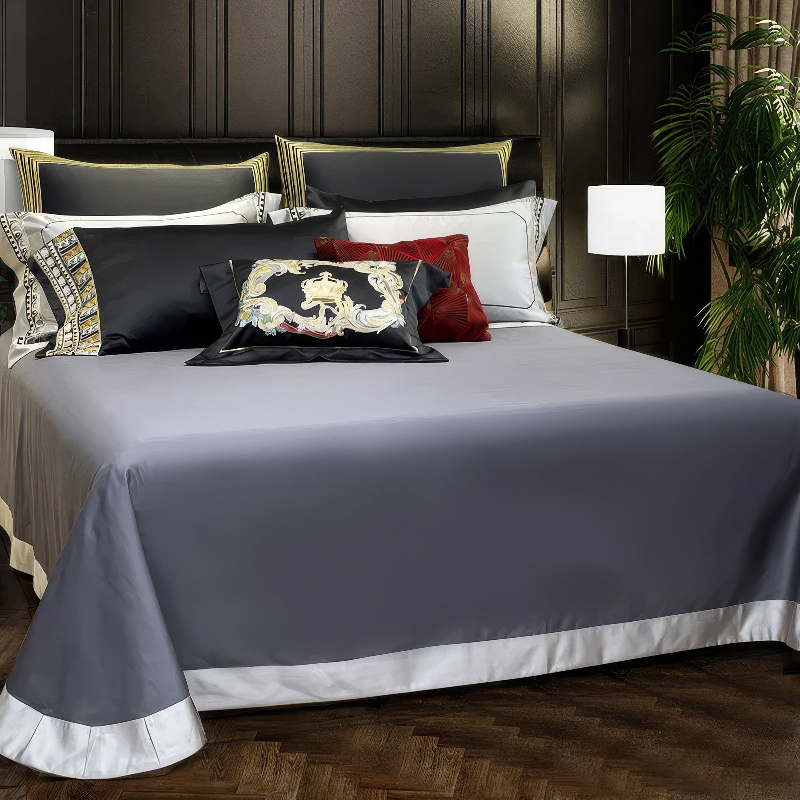 A sophisticated bedspread in slate gray with a contrasting white border, creating a refined and modern look. Accented by decorative pillows featuring intricate gold patterns and a bold red textured design, the bedding exudes luxury against a dark paneled wall and ambient lighting.

