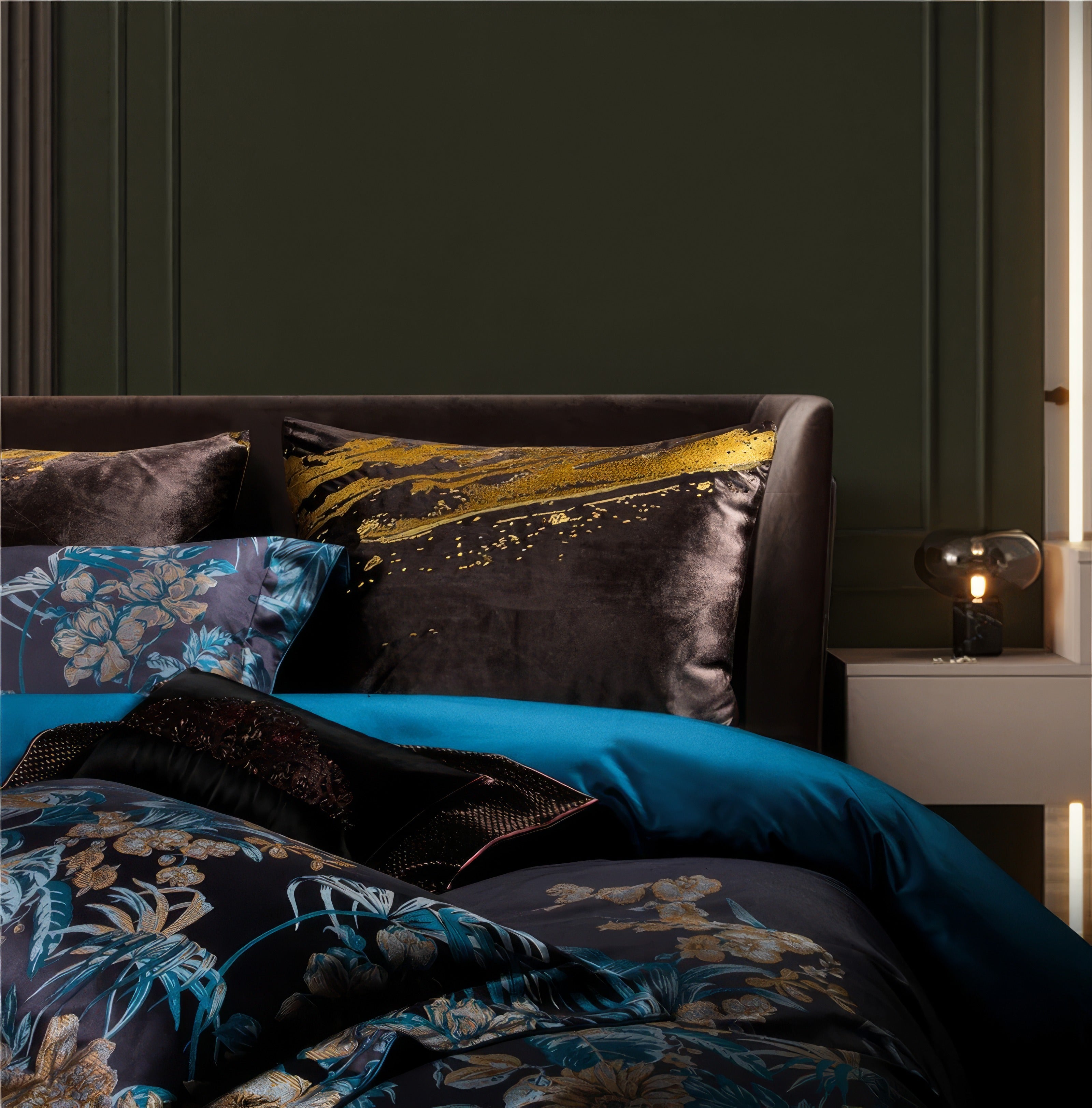 This close-up showcases the luxurious detailing of the bedding, focusing on the bold interplay between the black velvet pillow with gold accents and the vibrant floral patterns in blue and gold. The velvet pillow exudes opulence with its golden abstract streaks, adding a contemporary art-inspired touch to the overall aesthetic. The teal bedsheet beneath provides a striking contrast, enhancing the richness of the textures and colors. This arrangement perfectly blends modern luxury with timeless elegance, cre