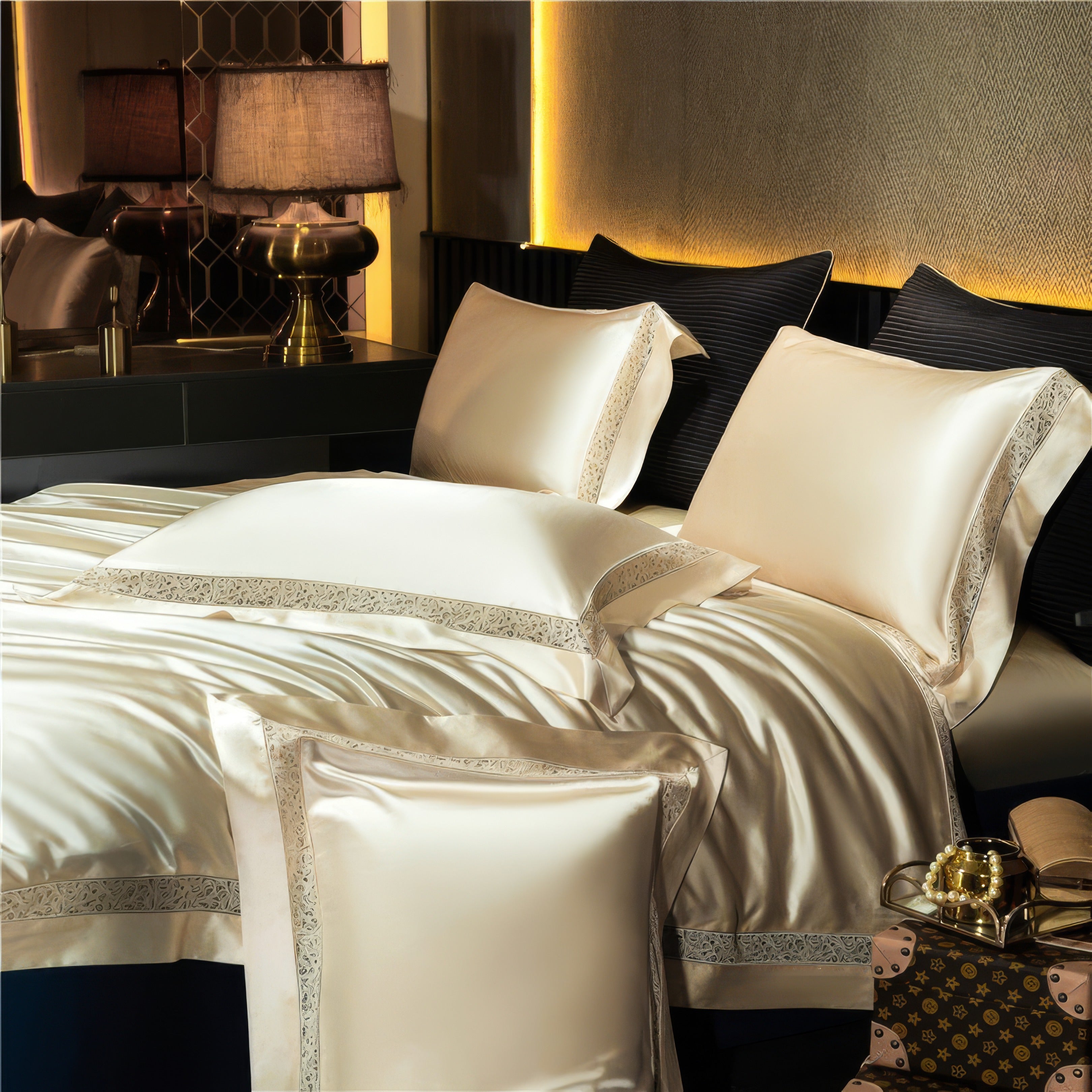 Close-up of a luxurious cream-colored bedding set featuring a silky finish and elegant embroidered trim on the pillowcases and duvet cover. The bed is adorned with plump pillows, contrasting black accent cushions, and a rich, illuminated backdrop. A bedside table with an ornate golden lamp and decorative accents adds sophistication, while a stylish designer travel case and accessories placed at the foot of the bed enhance the room’s opulent ambiance.