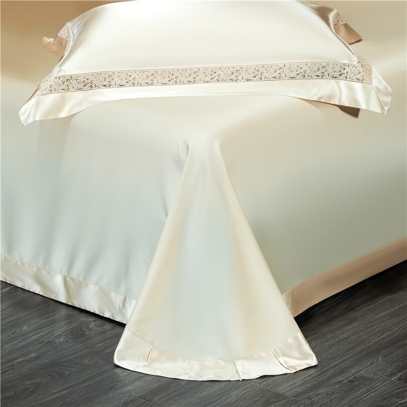 Elegant cream-colored bedspread draped over a bed, with intricate lace detailing along the edges of the pillowcase and hemline. The silky fabric flows smoothly, pooling gracefully onto the dark wooden floor, highlighting its luxurious texture and design. The minimalistic background accentuates the timeless sophistication of the bedding.