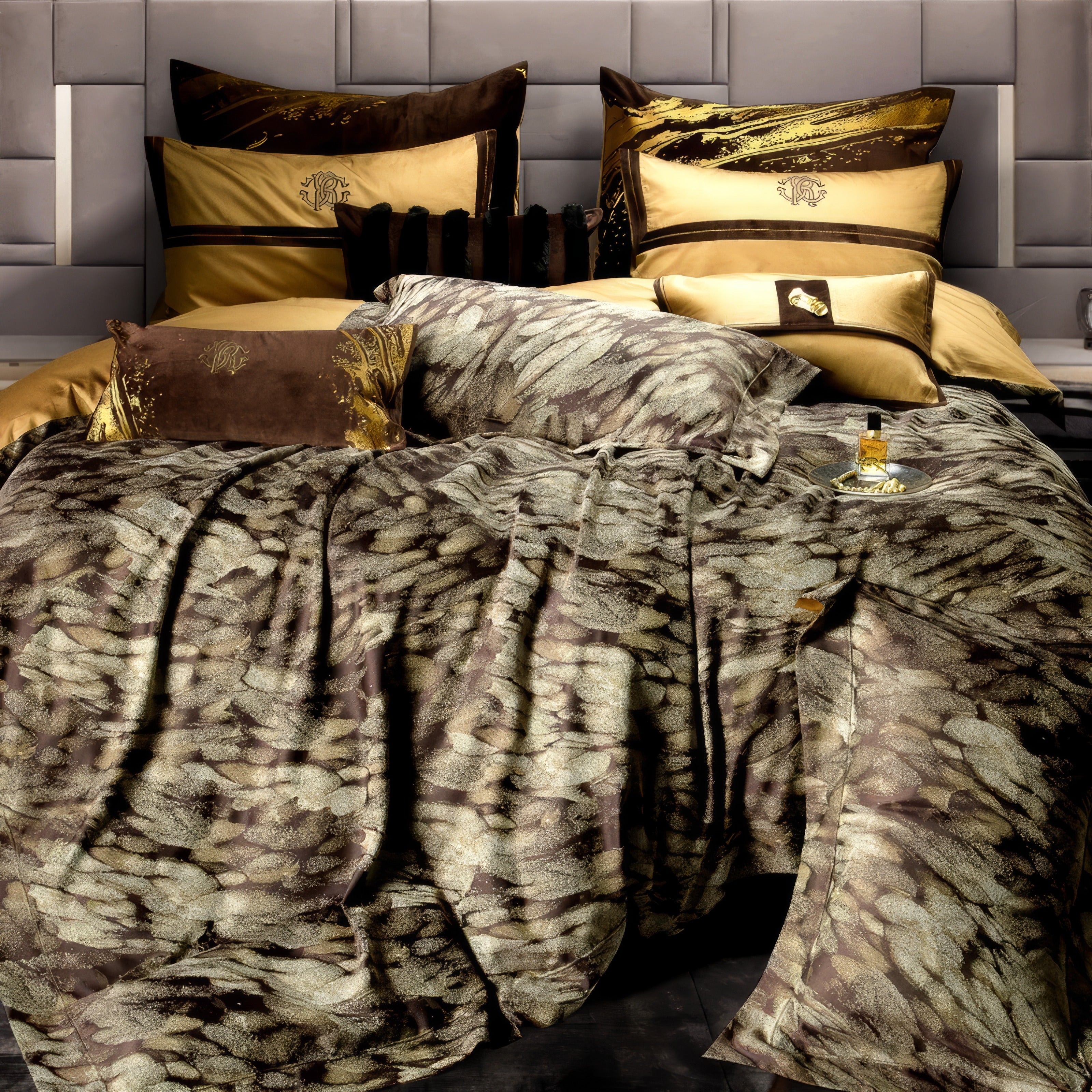 A close-up of a luxurious bedding set featuring a unique feather-inspired textured pattern in earthy brown and gold tones. The set includes a variety of decorative pillows with intricate embroidery and golden accents, adding layers of elegance and sophistication. The fabric's rich texture and sheen catch the light, emphasizing its opulent quality. A gold tray with a perfume bottle complements the bedding, enhancing the luxurious ambiance. The background showcases a soft grey upholstered headboard, creating 