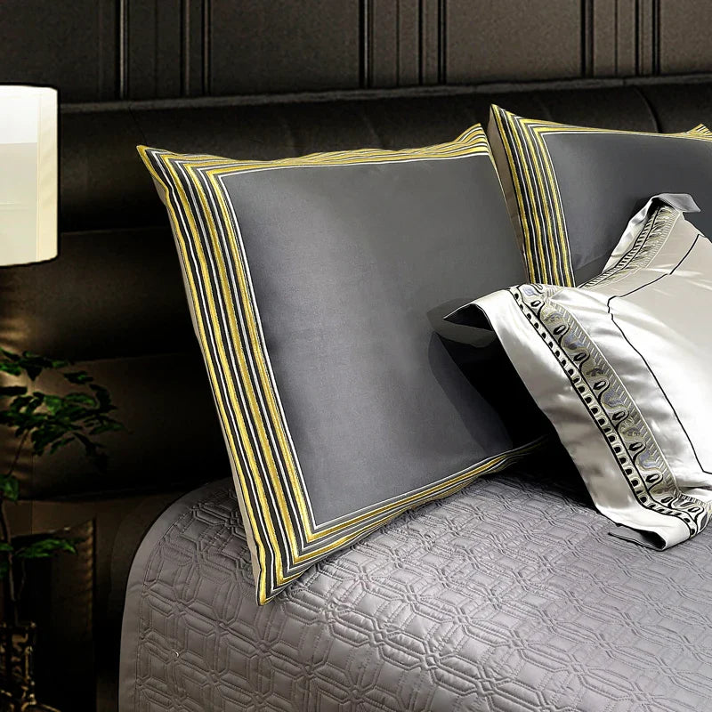 A close-up of a dark grey pillowcase bordered with a striking gold-striped trim, creating a modern and opulent look. The pillow sits on a quilted grey bedspread with a geometric pattern, complementing the sleek design. Adjacent is a contrasting white satin pillowcase adorned with a decorative black and silver embroidered border, enhancing the sophisticated ambiance of the bedding ensemble. The subtle lighting adds depth and highlights the luxurious textures and details.

